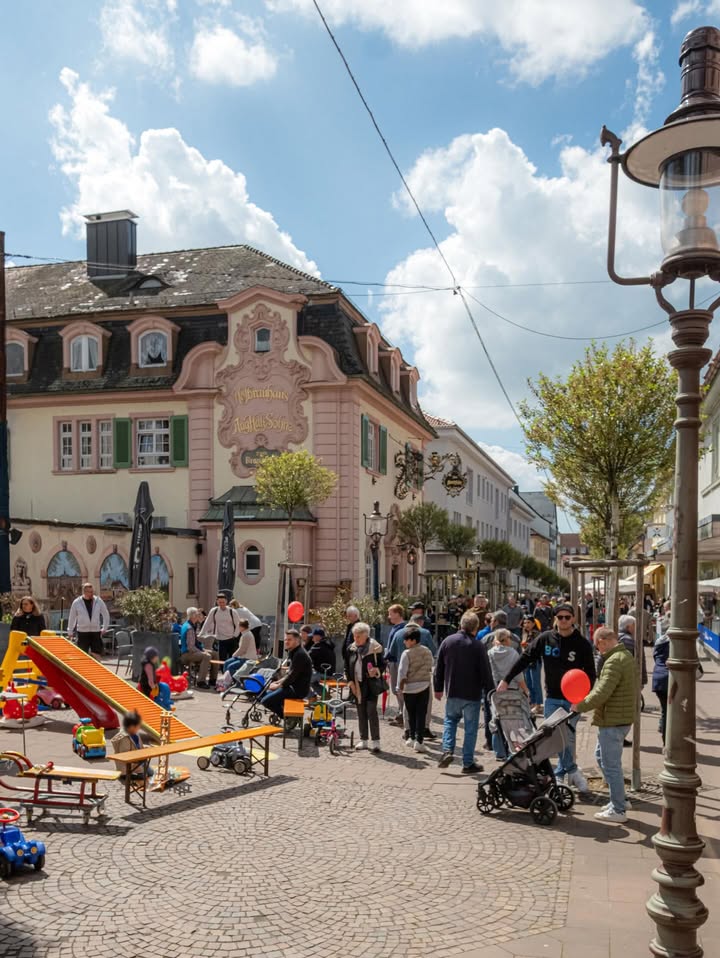 Dieser Alternativtext wurde KI-generiert: Ein Straßenszene mit einem historischen Gebäude im Hintergrund und Menschen, die sich auf einem Platz aufhalten. Im Vordergrund befindet sich ein Kinderspielplatz.