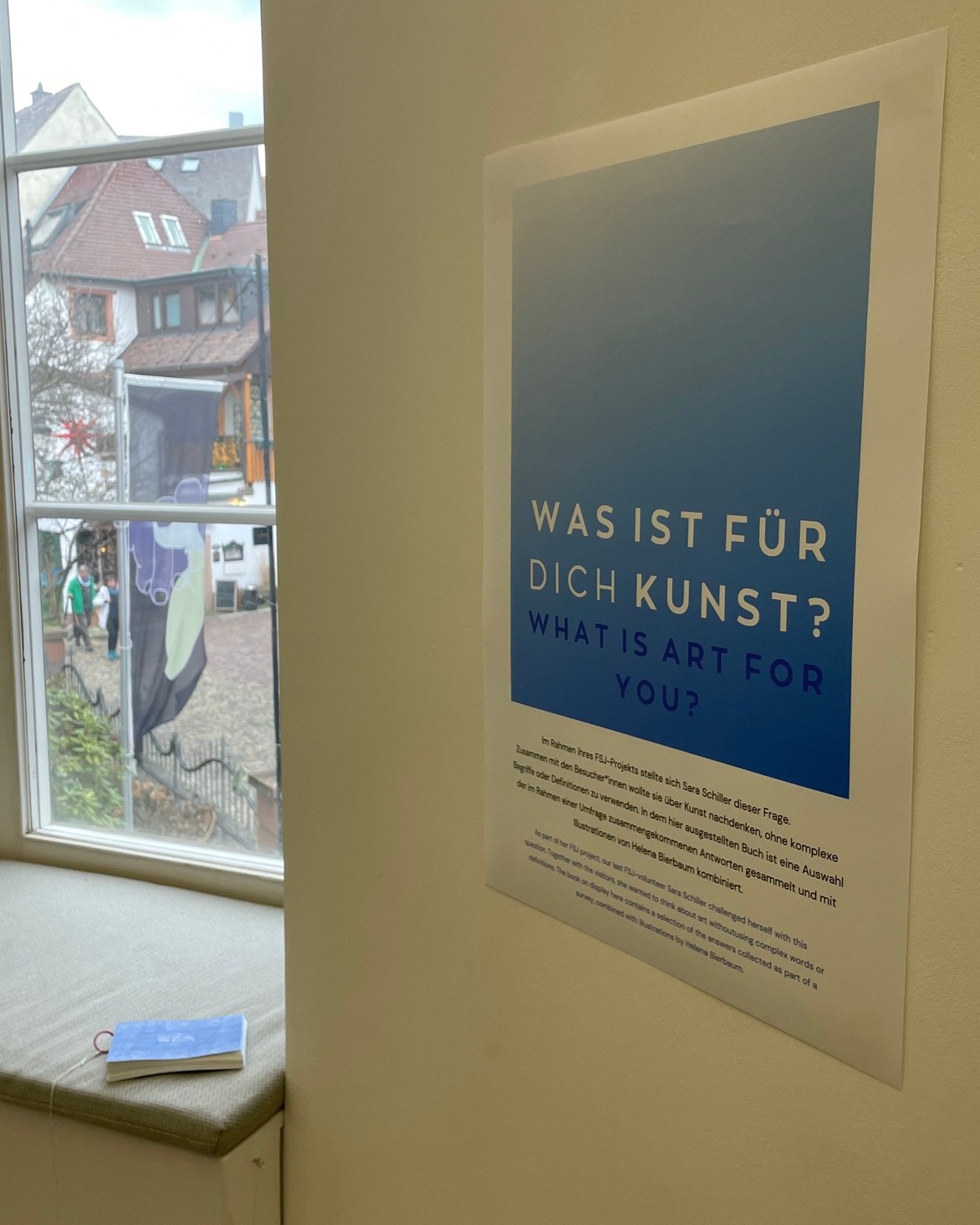 Dieser Alternativtext wurde KI-generiert: Ein Plakat mit Text in deutscher und englischer Sprache ist an einer Glaswand angebracht. Der Text beschäftigt sich mit der Frage, was Kunst für den Einzelnen bedeutet.