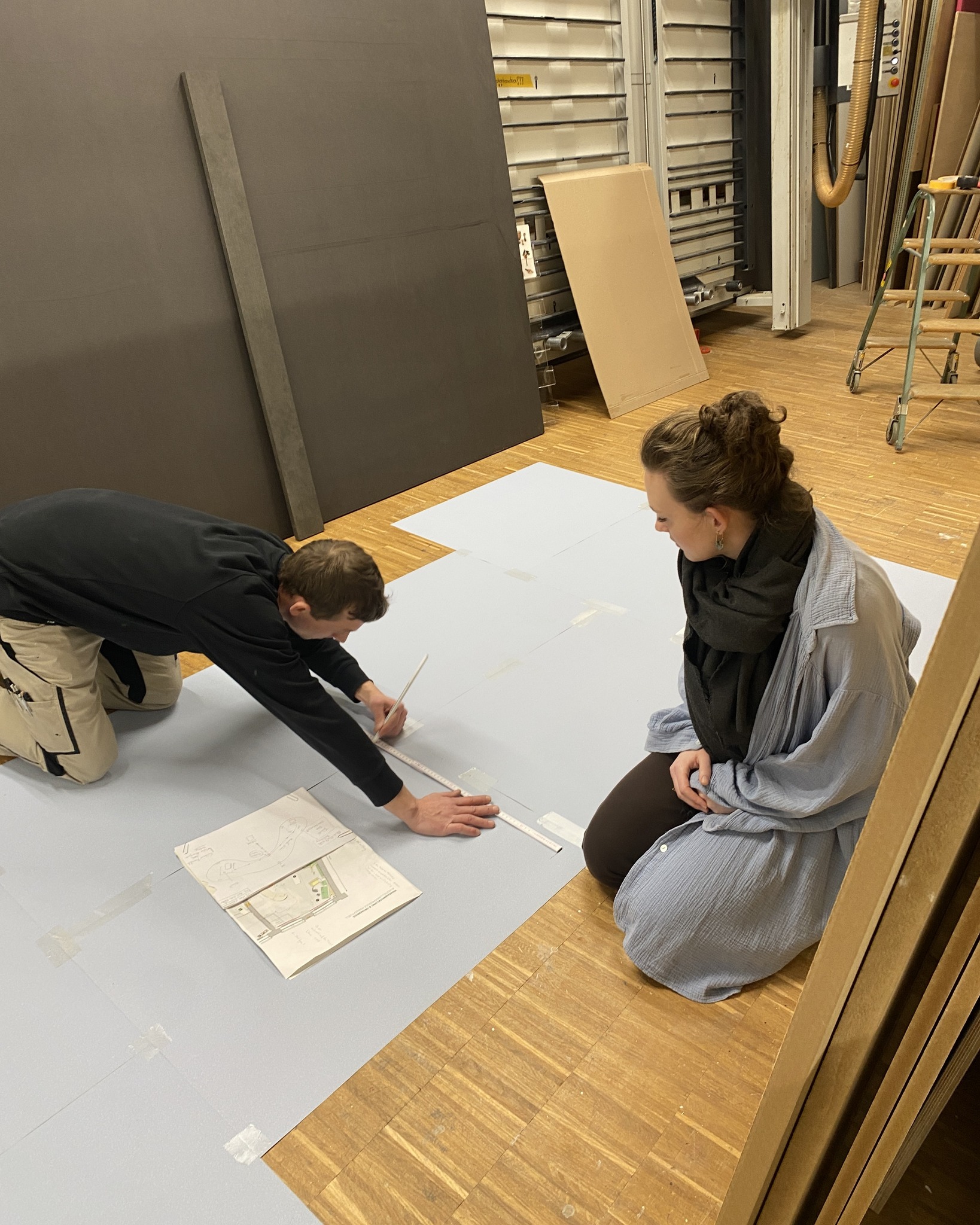 Dieser Alternativtext wurde KI-generiert: Zwei Personen sitzen oder knien auf dem Boden und betrachten Pläne auf einer großen Fläche. Im Hintergrund sind Regale und weitere Materialien zu sehen.