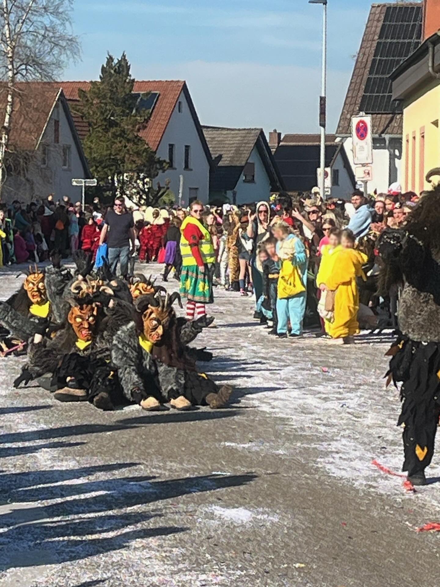 Dieser Alternativtext wurde KI-generiert: Eine Gruppe von Personen in farbenfrohen Kostümen steht auf einer Straße. Im Hintergrund sind Häuser und Bäume zu sehen.