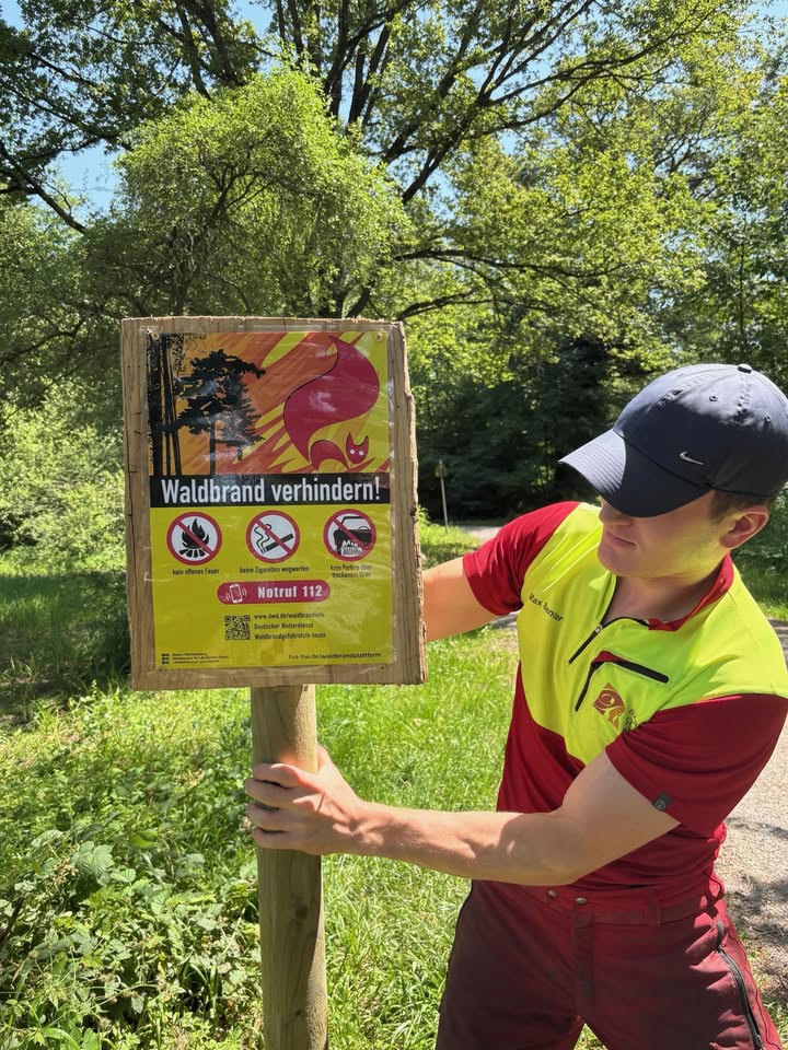 Eine Person in roter und gelber Arbeitskleidung hält einen Holzpfeiler mit einem Schild im Wald. Das Schild trägt die Aufschrift „Waldbrand verhindern!“, zeigt drei Verbotszeichen und die Notrufnummer 112. (KI-Alternativtext)