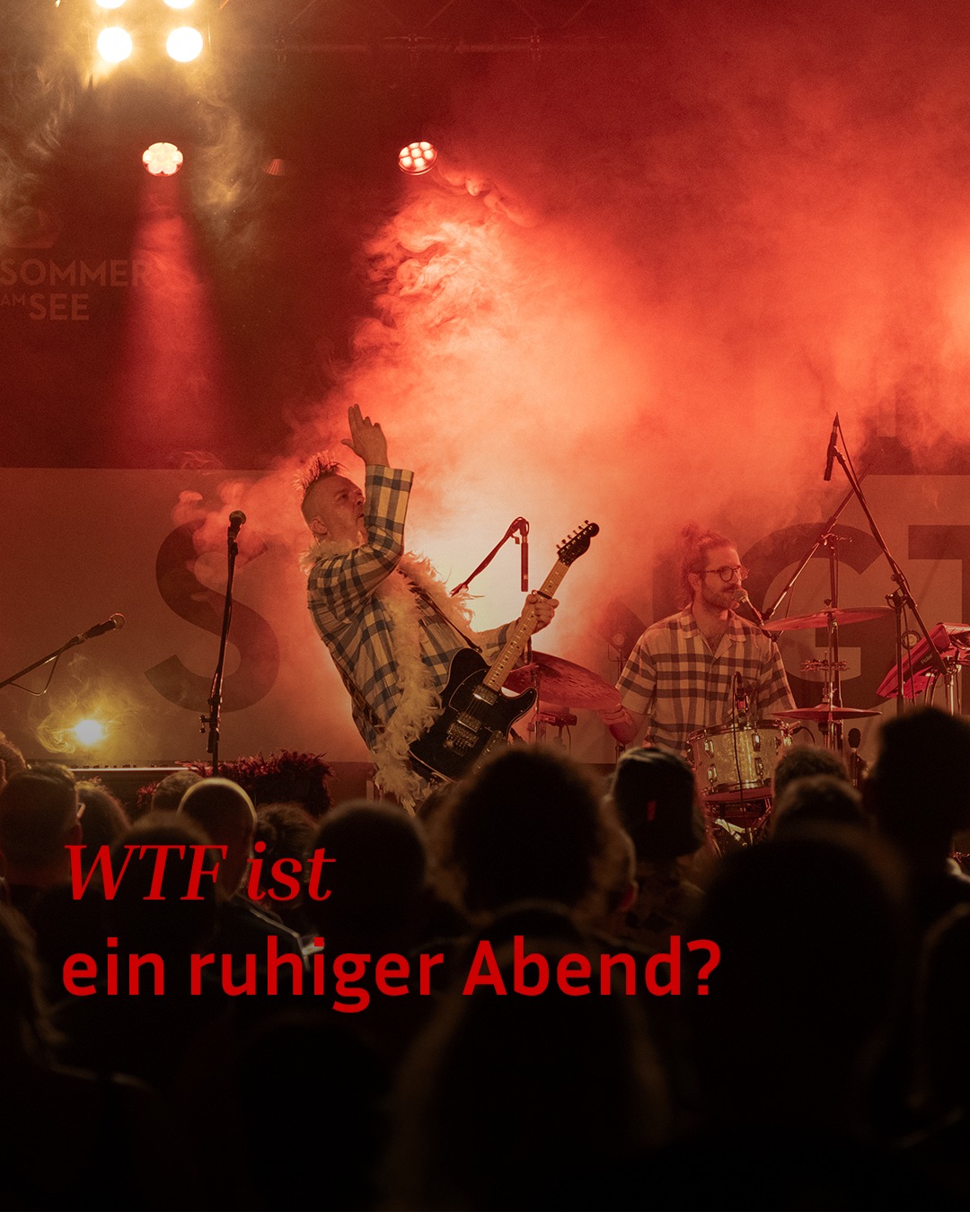 Zwei Personen spielen auf einer Bühne mit roter Beleuchtung und Raucheffekten Musik. Eine Person mit Federboa spielt E-Gitarre und hebt eine Hand, während die andere Schlagzeug spielt. Im Vordergrund sind Silhouetten des Publikums zu sehen, darüber steht der rote Text "WTF ist ein ruhiger Abend?". (KI-Alternativtext)