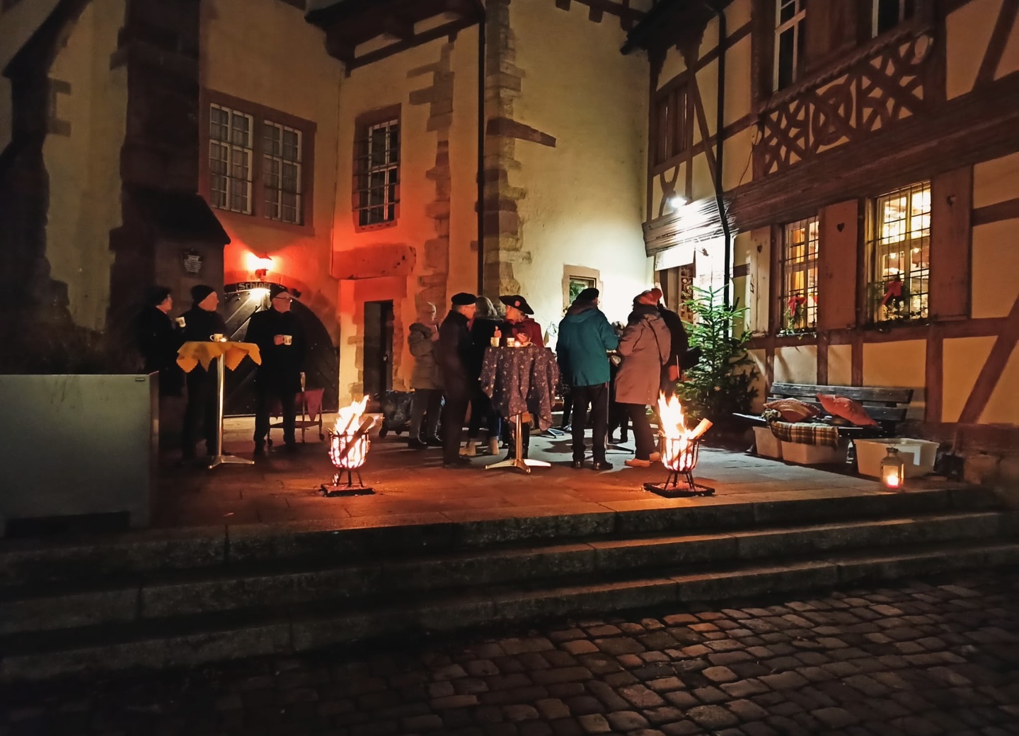 Menschen stehen an einem Abend vor einem alten Gebäude an kleinen Feuern zusammen. Es ist gemütlich und warm beleuchtet.