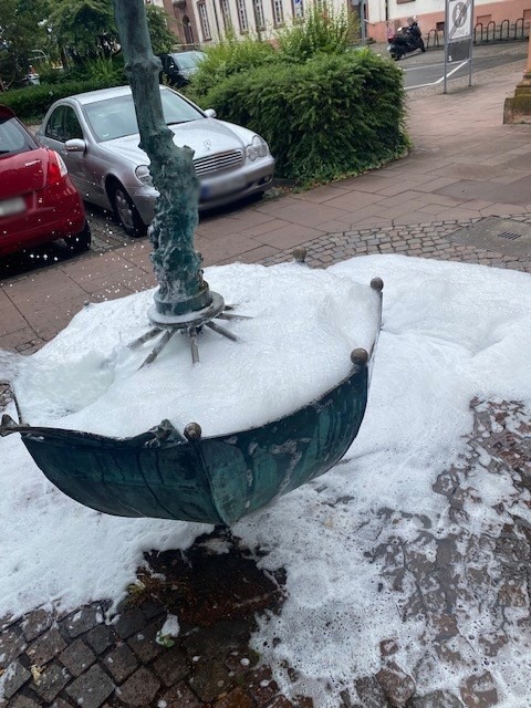 Eine grüne Skulptur in Form eines umgedrehten Regenschirms, gefüllt mit weißem Schaum, steht auf einem gepflasterten Platz. Im Hintergrund sind geparkte Autos, Büsche und ein Gebäude zu sehen. (KI-Alternativtext)