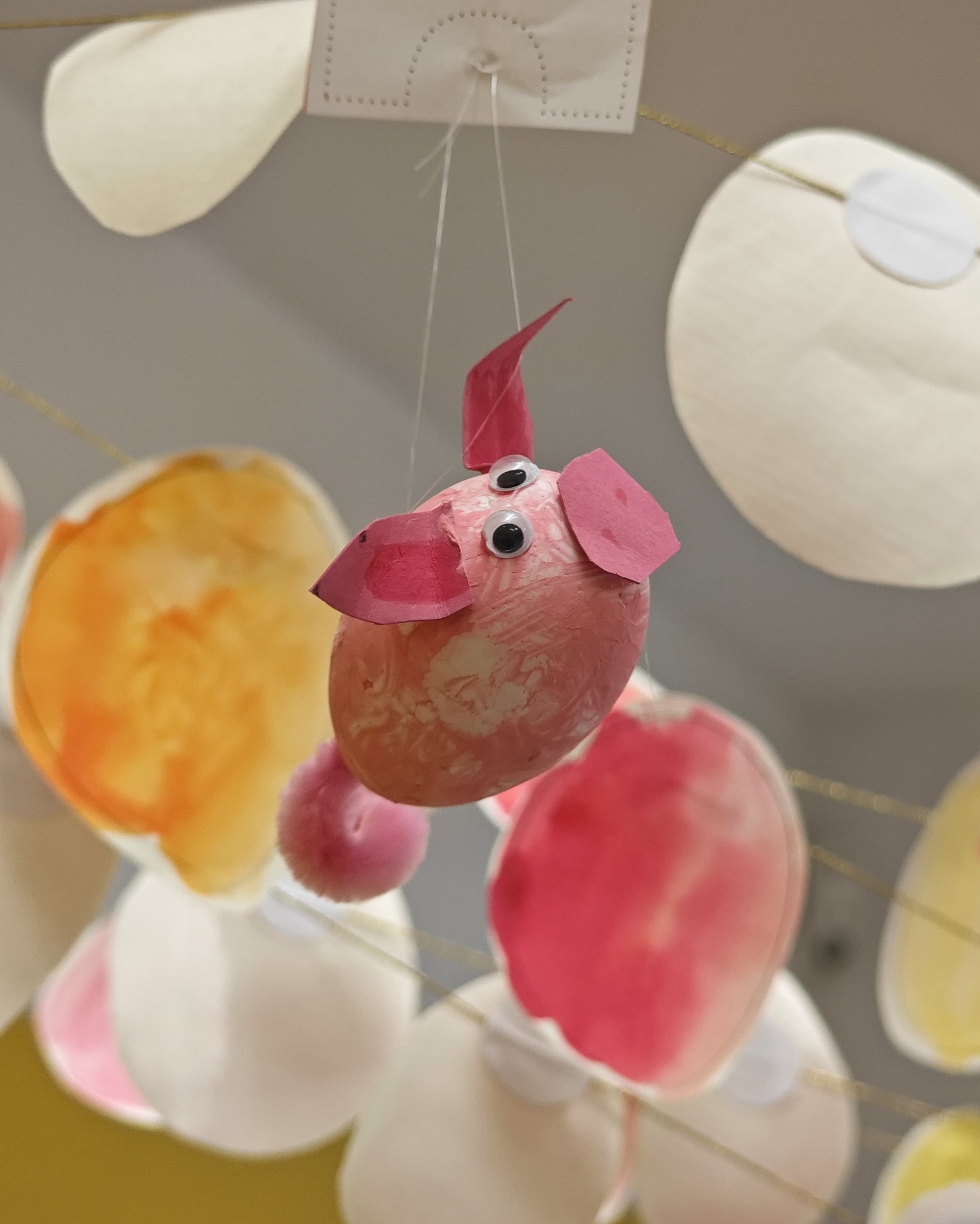 Dieser Alternativtext wurde KI-generiert: Eine farbenfrohe, handgefertigte Dekoration hängt an einer Schnur.  Es zeigt mehrere Formen, darunter ein rosafarbenes Objekt mit angedeuteten tierischen Merkmalen.