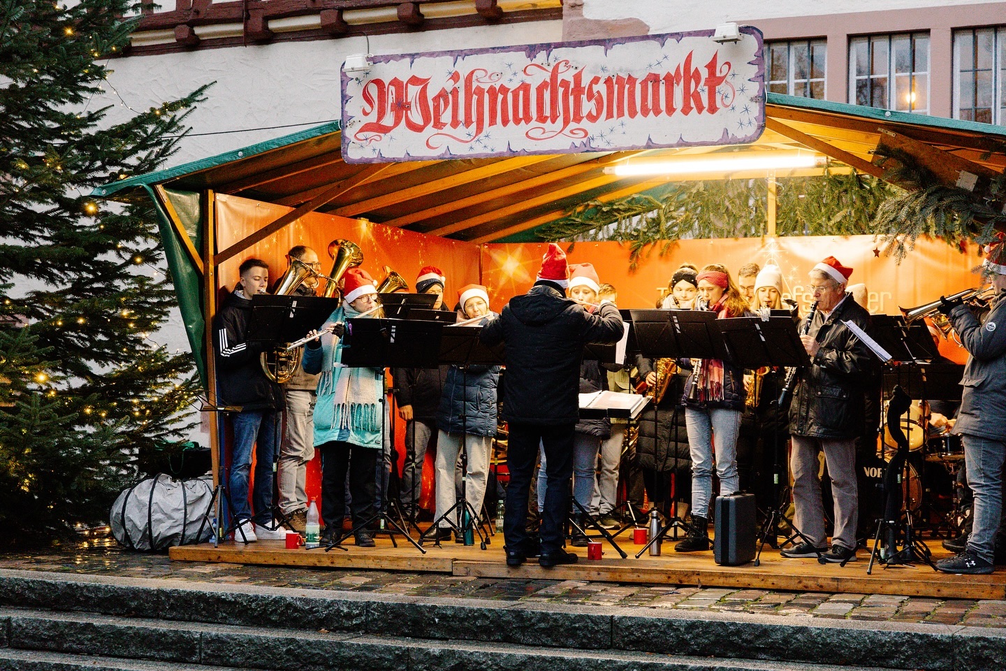 Eine Musikgruppe spielt auf einem Weihnachtsmarkt. Die Musiker tragen rote Mützen und spielen verschiedene Blasinstrumente.
