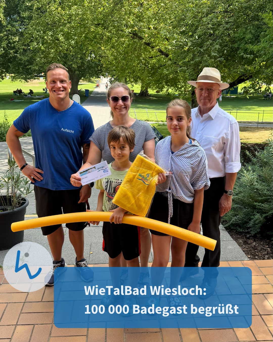 Fünf Personen stehen im Freien vor Bäumen. Eine Person trägt ein blaues T-Shirt mit der Aufschrift „Aufsicht“. Ein Kind hält eine gelbe Pool-Noodle, eine weitere Person ein gelbes Handtuch. Ein Textfeld unten lautet: „WieTalBad Wiesloch: 100 000 Badegast begrüßt“. (KI-Alternativtext)
