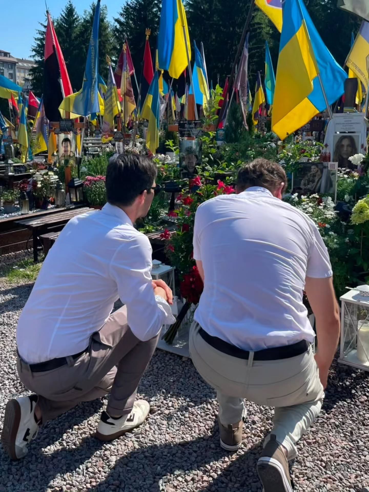 Zwei Personen in weißen Hemden und hellen Hosen knien auf einem Kiesweg vor Gräbern, die mit zahlreichen ukrainischen Flaggen, Blumen und Porträtfotos geschmückt sind. (KI-Alternativtext)