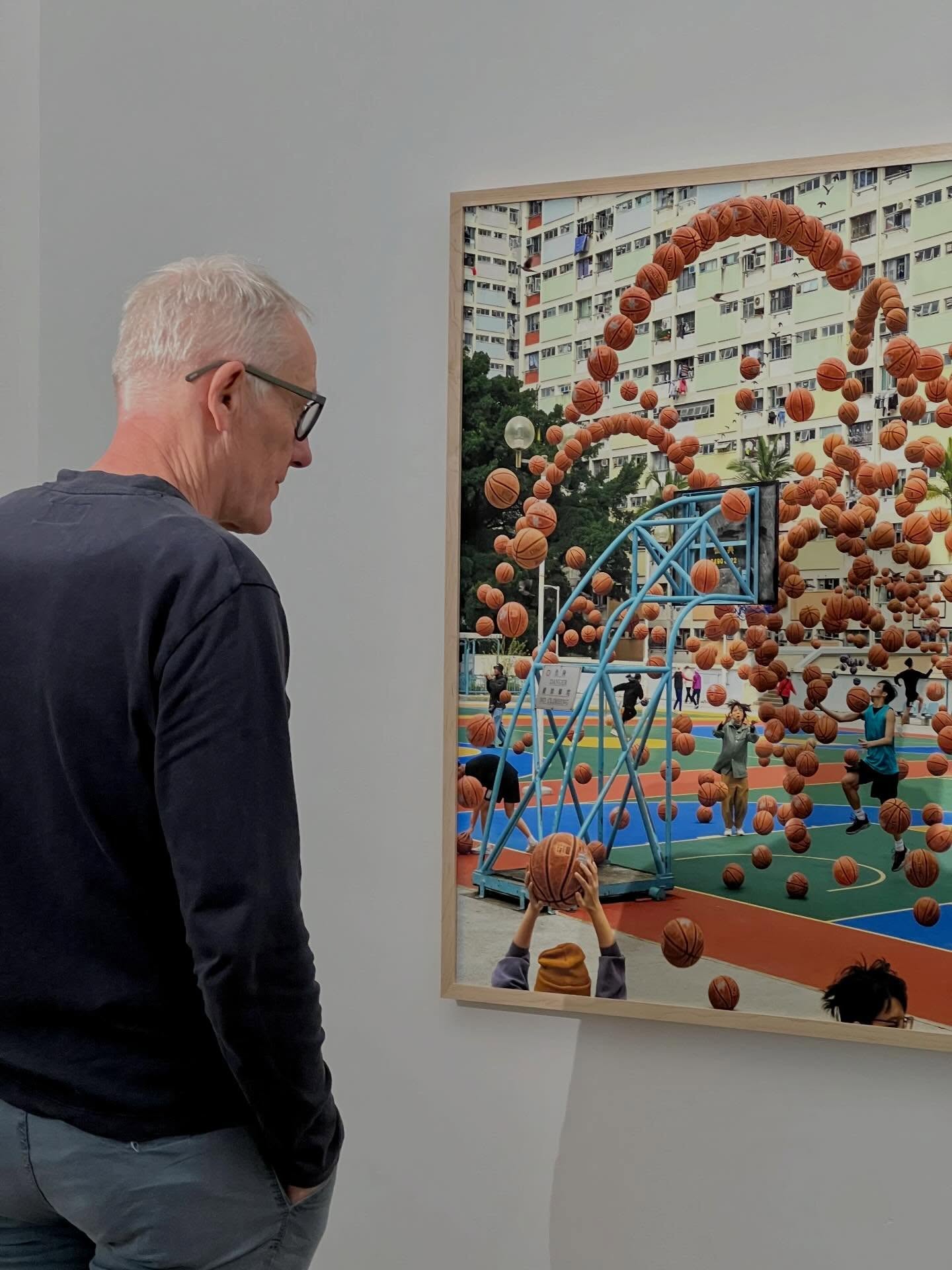 Dieser Alternativtext wurde KI-generiert: Ein Mann betrachtet ein farbenfrohes Kunstwerk, das eine Basketballszene mit zahlreichen Bällen zeigt. Das Bild hängt an einer hellen Wand.