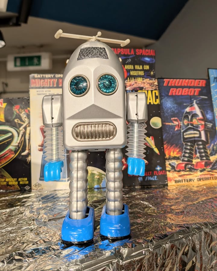 Dieser Alternativtext wurde KI-generiert: Ein vintage Blechroboter steht vor Regalen mit Spielzeugverpackungen. Das Bild ist farbig und zeigt einen Retro-Look mit Sci-Fi-Elementen.