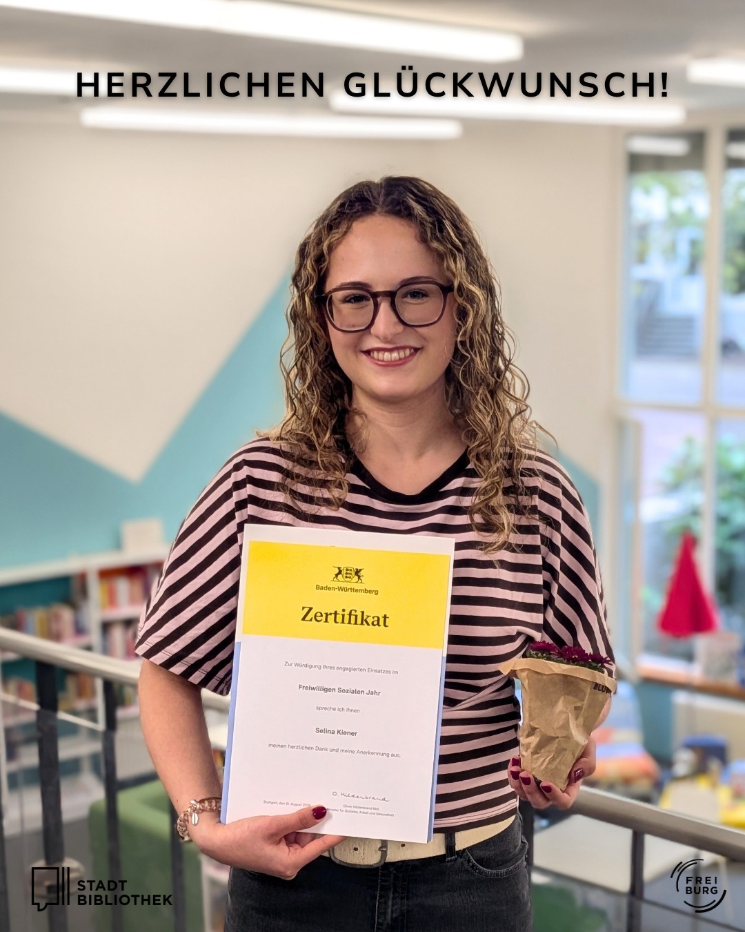 Eine Person mit Brille und gestreiftem Oberteil hält ein Zertifikat für das Freiwillige Soziale Jahr sowie einen kleinen Blumenstrauß. Oben im Bild steht der Text „HERZLICHEN GLÜCKWUNSCH!“. Unten links und rechts sind die Logos der Stadtbibliothek und von Freiburg zu sehen. (KI-Alternativtext)