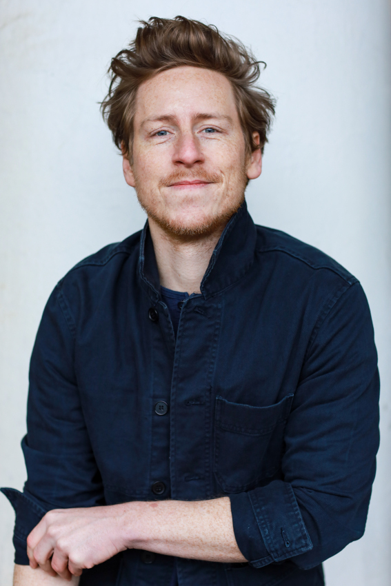 Dieser Alternativtext wurde KI-generiert: Porträt einer Person mit hellen Haaren und Bart. Die Person trägt ein dunkles Oberteil und blickt in die Kamera.