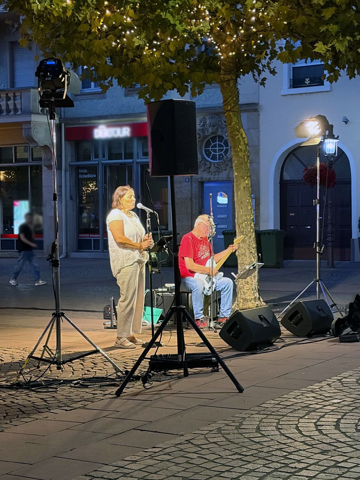 Zwei Personen geben eine Straßenmusikdarbietung bei Nacht auf einem gepflasterten Platz. Eine Person steht und singt in ein Mikrofon, während die andere sitzend eine E-Gitarre spielt. Um sie herum stehen Lautsprecher, Mikrofonständer und Beleuchtungstechnik, im Hintergrund ist ein Geschäft mit dem Schild „DETOUR“ zu sehen. (KI-Alternativtext)