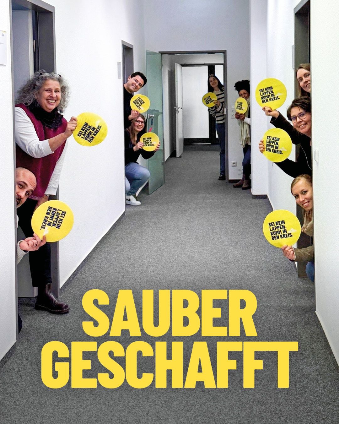 Dieser Alternativtext wurde KI-generiert: Eine Gruppe von Menschen steht in einem Flur und hält gelbe Plakate mit schwarzem Text. Im Vordergrund steht ein großes, farbiges Schild mit der Aufschrift "SAUBER GESCHAFFT".