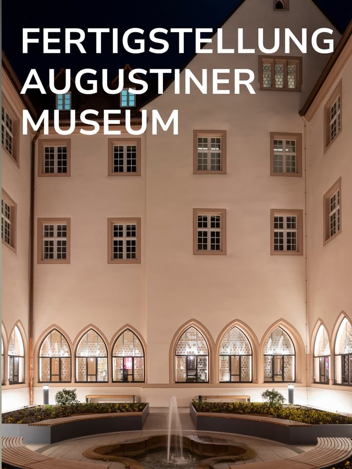 Große weiße Schrift oben: FERTIGSTELLUNG AUGUSTINER MUSEUM. Im Hintergrund ein beleuchtetes Gebäude mit hellen Fassaden, rechteckigen Fenstern im Obergeschoss und hohen Bogenfenstern im Erdgeschoss. Im Vordergrund ein Innenhof mit einem zentralen Brunnen, umgeben von Pflanzbeeten und Bänken. (KI-Alternativtext)