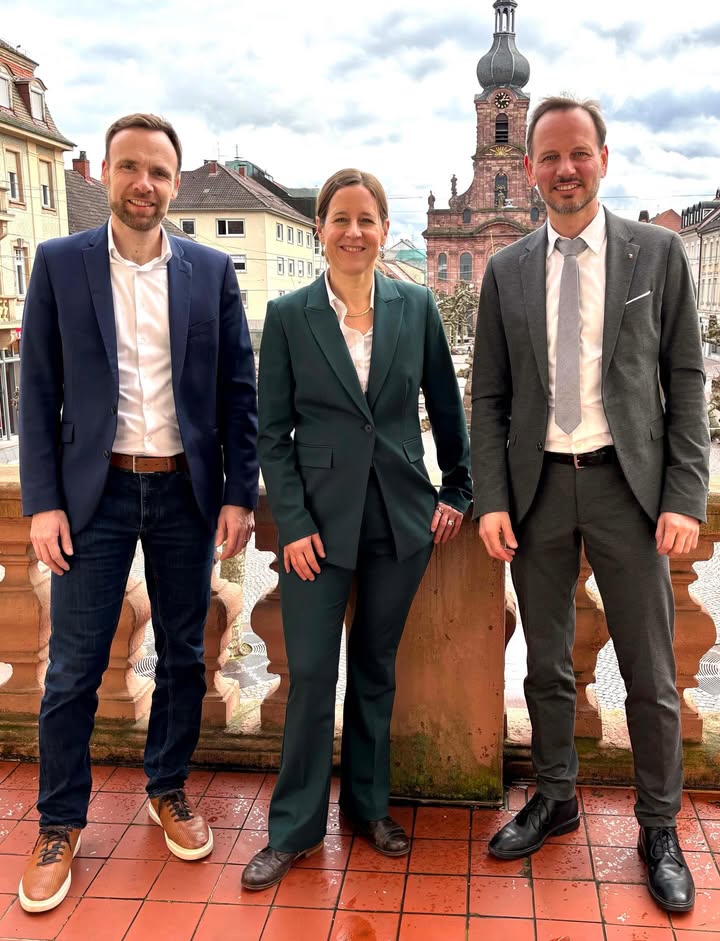 Dieser Alternativtext wurde KI-generiert: Drei Personen in formeller Kleidung stehen im Freien vor einer historischen Stadtkulisse. Im Hintergrund ist ein Kirchturm mit einer Uhr zu sehen.