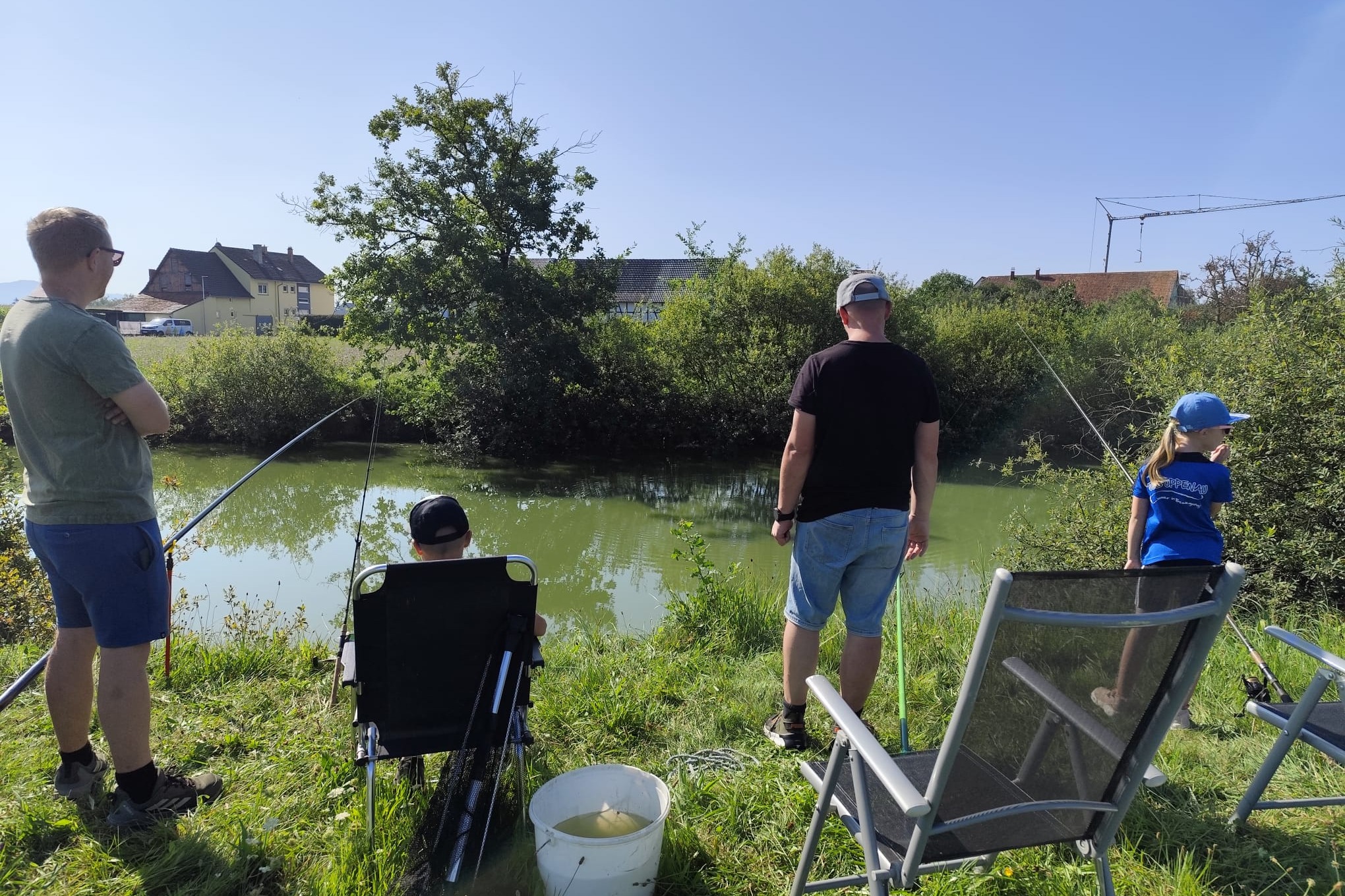 Ein farbiges Bild zeigt Personen beim Angeln an einem Gewässer mit üppiger Vegetation. Im Hintergrund sind Häuser und Bäume zu sehen.