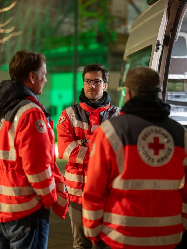 Dieser Alternativtext wurde KI-generiert: Drei Personen in heller Warnweste stehen im Freien neben einem Fahrzeug. Im Hintergrund ist grünes Licht zu sehen.