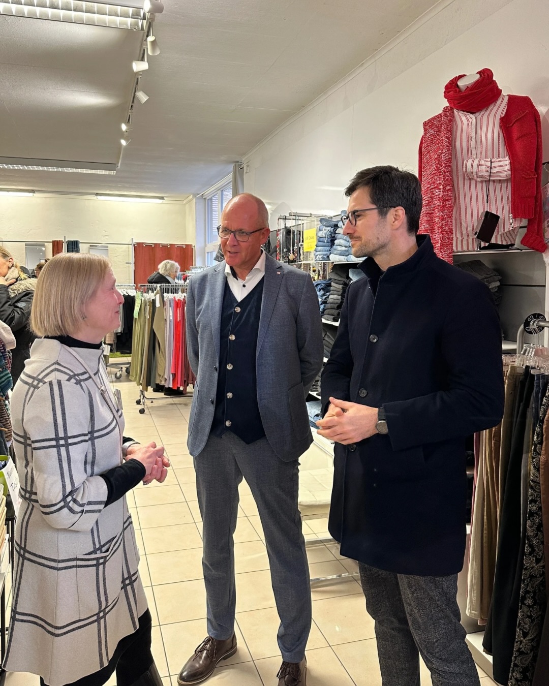 Dieser Alternativtext wurde KI-generiert: Mehrere Personen stehen in einem Geschäft mit Kleidung und unterhalten sich. Im Hintergrund hängen Kleidungsstücke auf Bügeln.