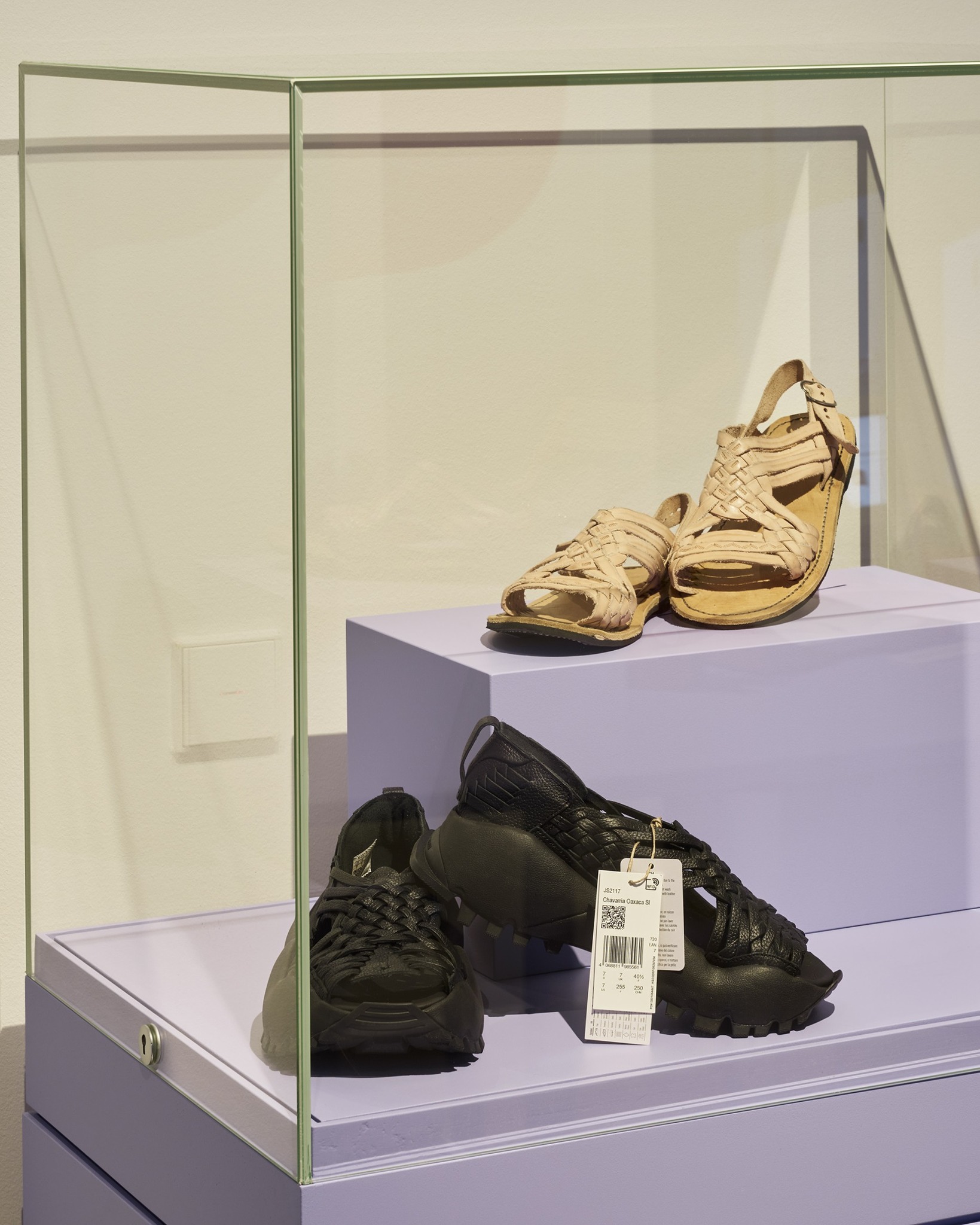 Glasvitrine mit lila Regalen, die zwei Paar Schuhe präsentiert. Oben stehen beige, geflochtene Sandalen. Unten liegen schwarze Sneaker mit dicken Sohlen, an denen ein weißes Etikett mit der Aufschrift „JS2117 Chavarría Oaxaca SI“ hängt. (KI-Alternativtext)