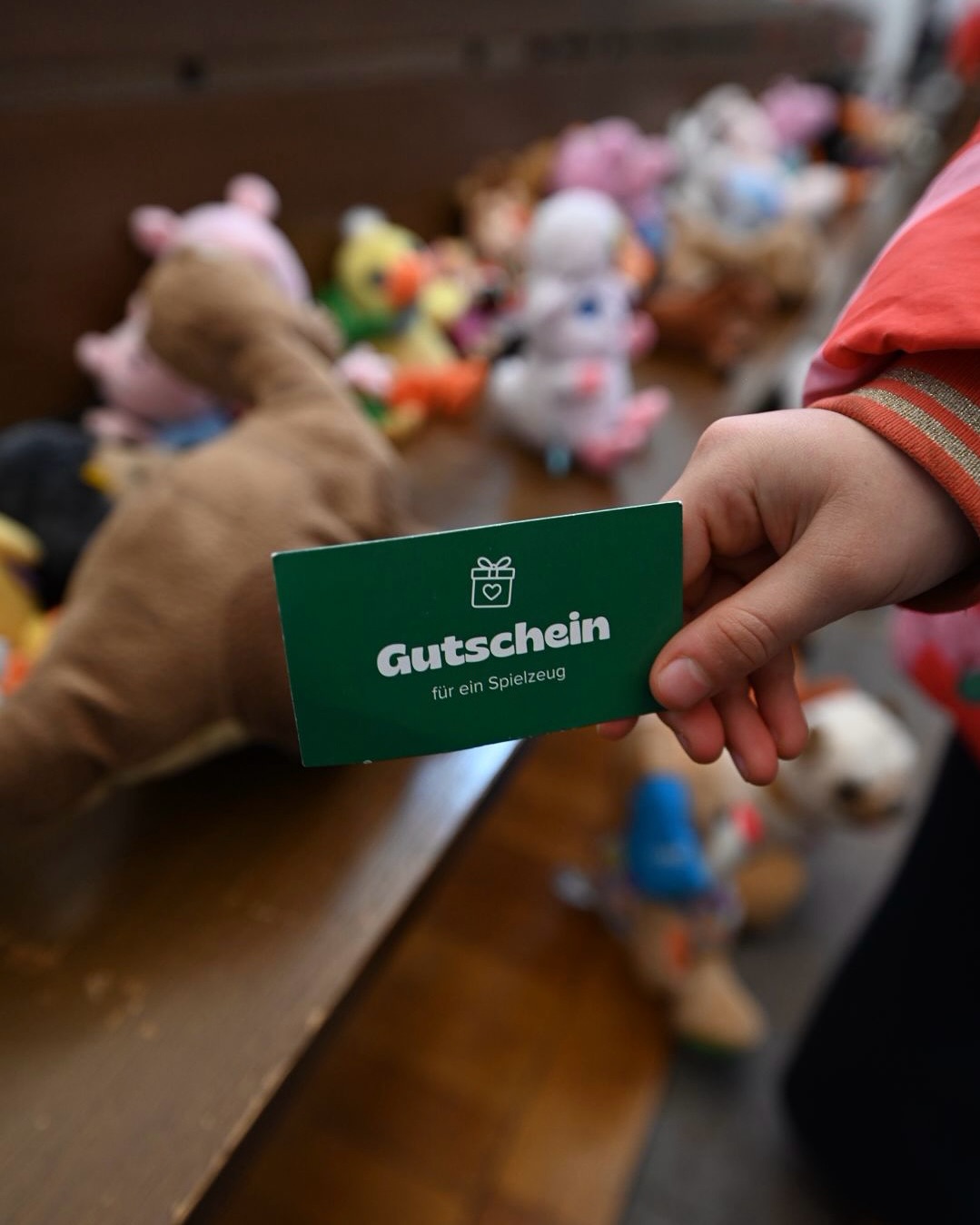 Dieser Alternativtext wurde KI-generiert: Eine Hand hält eine grüne Karte mit einem Geschenk-Symbol und der Aufschrift "Gutschein für ein Spielzeug". Im Hintergrund sind bunte Plüschtiere zu sehen.