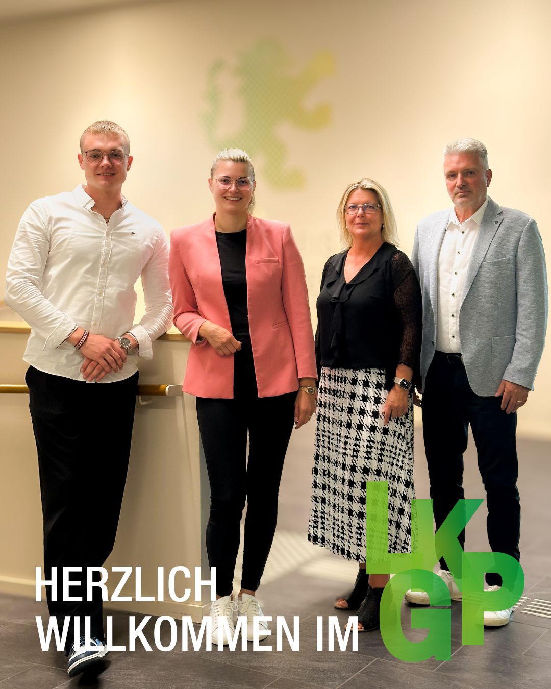 Vier Personen stehen nebeneinander in einem hellen Raum. Von links nach rechts tragen sie ein weißes Hemd, eine rosa Jacke, ein schwarzes Oberteil mit kariertem Rock und ein graues Sakko. Im Vordergrund ist der Text „HERZLICH WILLKOMMEN IM LKGP“ eingeblendet. (KI-Alternativtext)
