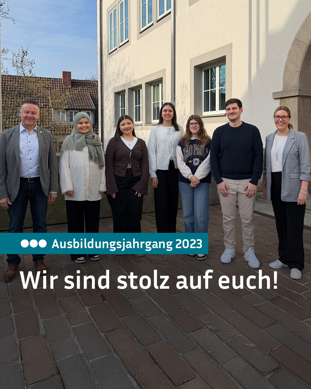 Sieben Personen stehen lächelnd vor einem Gebäude. Ein Textfeld unten links zeigt „Ausbildungsjahrgang 2023“, darunter steht „Wir sind stolz auf euch!“. Eine Person trägt eine Kopfbedeckung. (KI-Alternativtext)