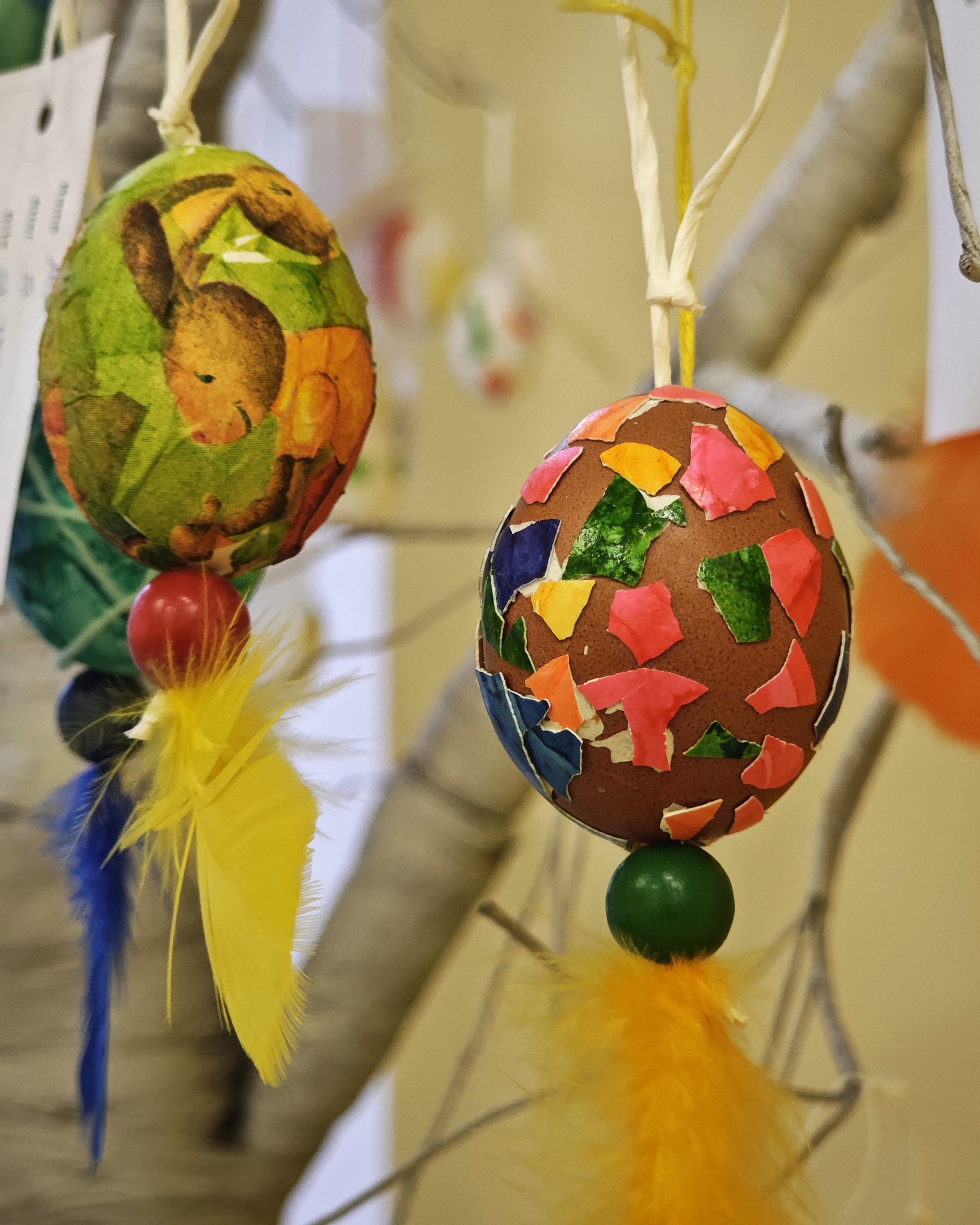Dieser Alternativtext wurde KI-generiert: Zwei verzierte Eier hängen an Fäden vor einem unscharfen Hintergrund. Die Eier sind mit bunten Materialien beklebt und haben farbige Federdekorationen.