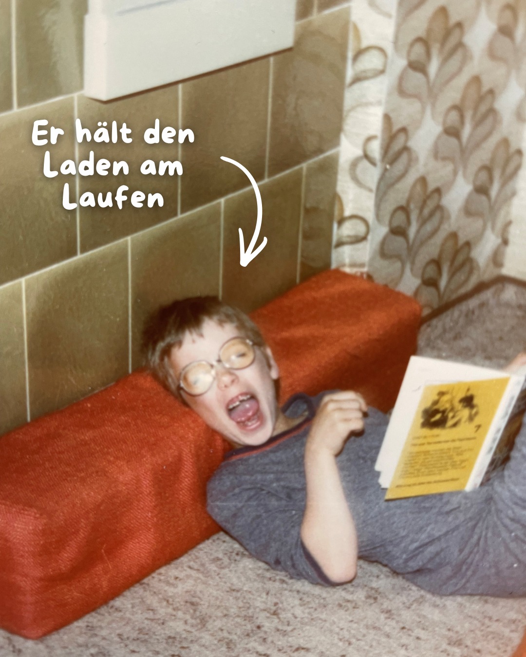 Dieser Alternativtext wurde KI-generiert: Ein Schwarz-Weiß-Foto zeigt eine Person, die auf einem roten Polster liegt und ein Buch hält. Im Hintergrund sind Fliesen an einer Wand und ein Vorhang zu sehen.