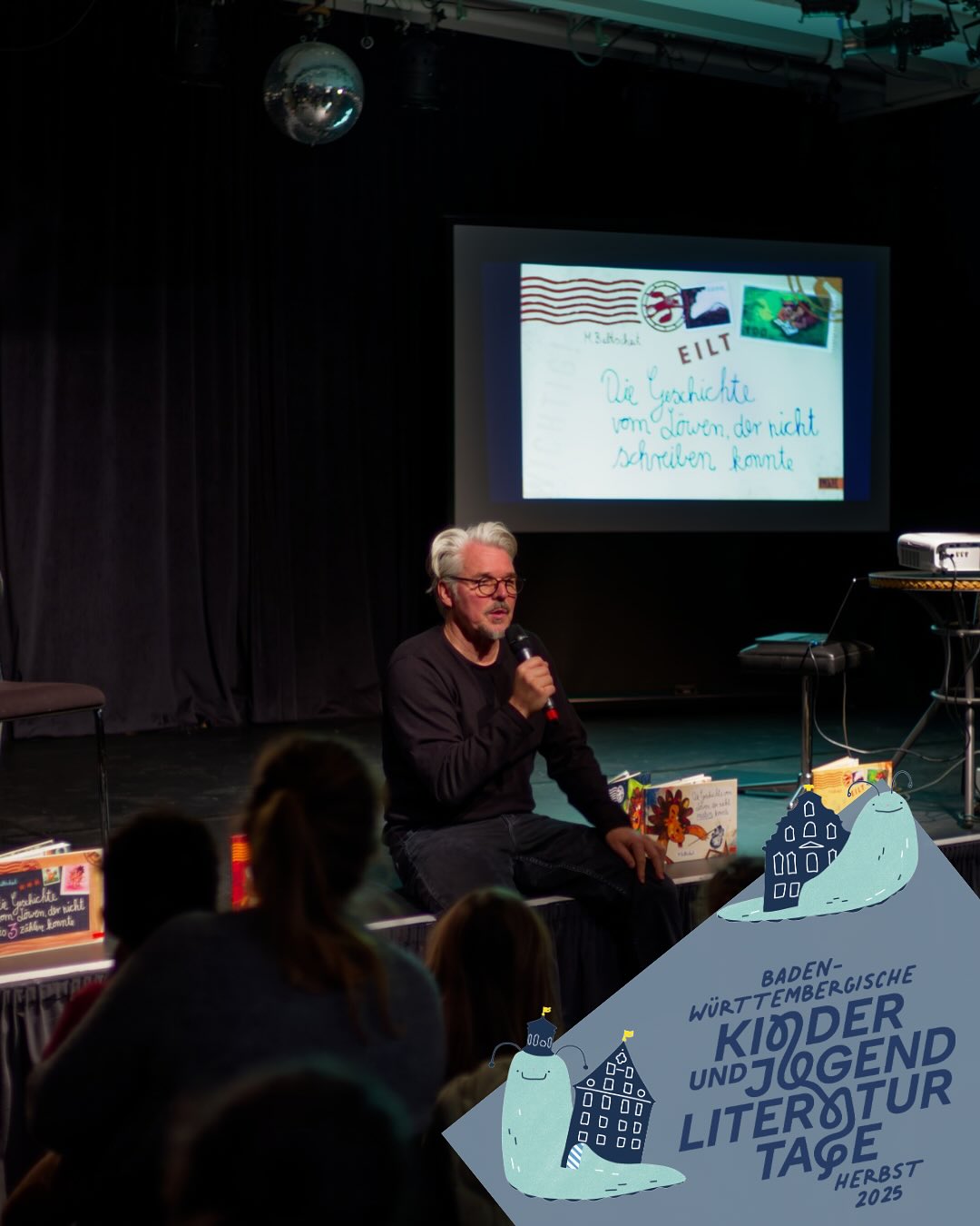 Dieser Alternativtext wurde KI-generiert: Eine Person spricht in einen Mikrofon vor einer Projektion und einem Publikum. Dekorationen und ein Banner mit Text sind ebenfalls im Bild zu sehen.