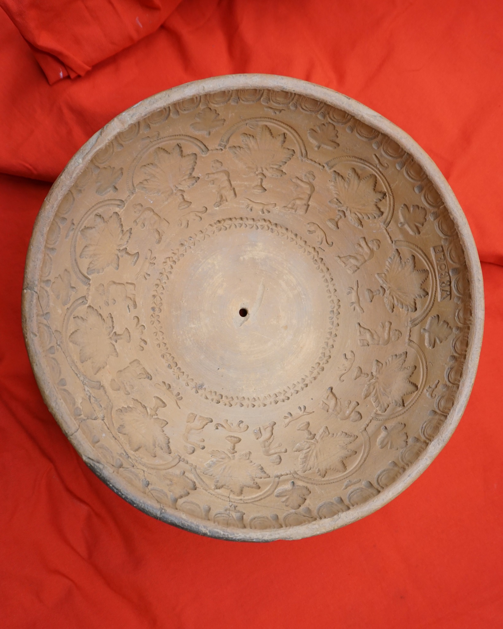 Der Boden einer beige gefärbten Keramikschale mit eingravierten Mustern aus Blättern und kleinen Figuren liegt auf rotem Stoff. In der Mitte befindet sich ein kleines Loch, am Rand ist der Schriftzug "VRBOLE" eingestempelt. (KI-Alternativtext)