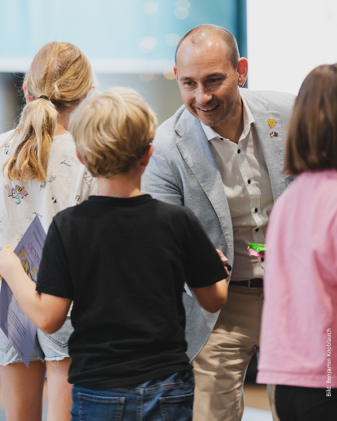 Ein Erwachsener in grauem Sakko lächelt drei Kindern zu, die ihm den Rücken zukehren; eines hält ein Blatt Papier mit der Aufschrift „ORTYP“. (KI-Alternativtext)