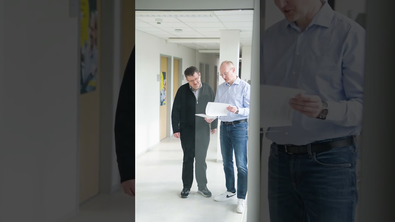 Dieser Alternativtext wurde KI-generiert: Zwei Personen stehen in einem Flur und betrachten ein Blatt Papier. Das Bild ist farbig und scheint in einem Innenraum aufgenommen worden zu sein.