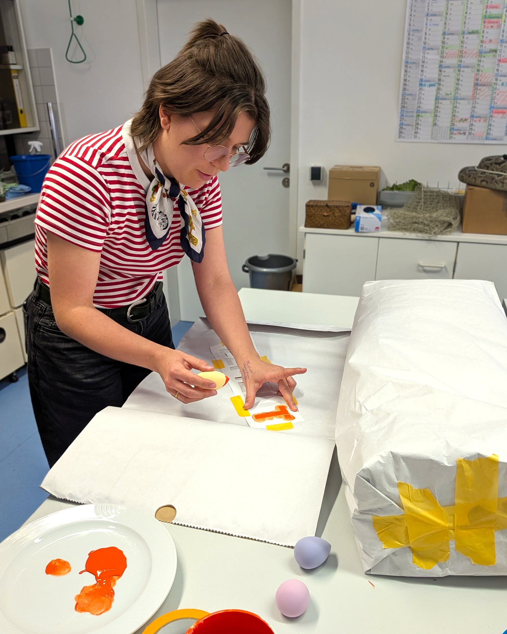 Eine Person in gestreiftem Shirt und Kopfbedeckung bemalt ein Ei mit orangefarbener Farbe, die mit einem Schablonenstempel auf Papier aufgetragen wird. Auf dem Tisch liegen weitere bunte Eier, Farbe auf einem Teller und ein mit Papier und Klebeband verpacktes Objekt. (KI-Alternativtext)