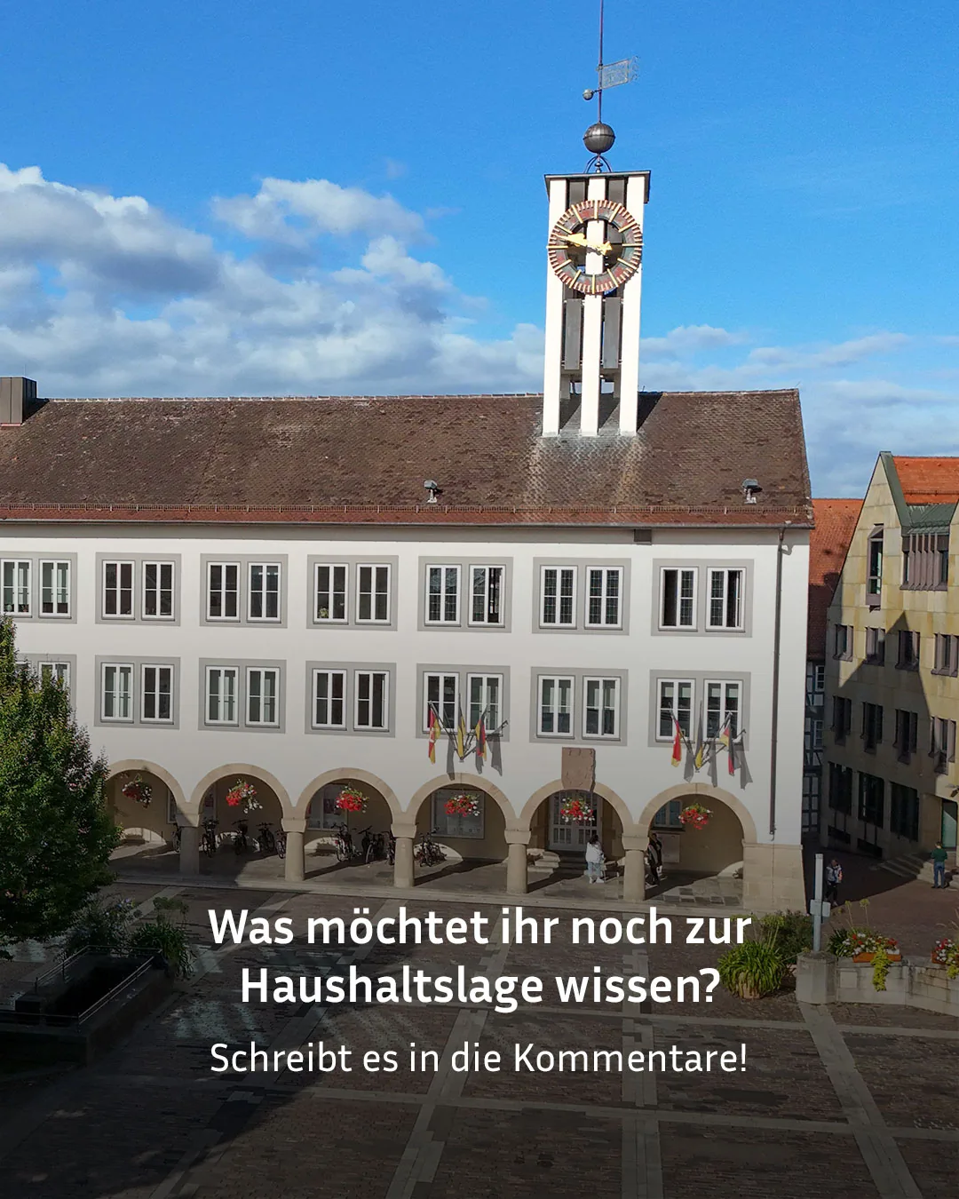 Ein weißes Gebäude mit einem hohen Uhrturm und Windfahne steht über einem gepflasterten Platz. Im Erdgeschoss befinden sich Bogenreihen mit Blumenkörben, darunter stehen Fahrräder und einige Personen gehen vor dem Eingang. Mehrere Flaggen hängen an den Fenstern des Obergeschosses. Der Text "Was möchtet ihr noch zur Haushaltsslage wissen? Schreibt es in die Kommentare!" ist am unteren Bildrand eingeblendet. (KI-Alternativtext)