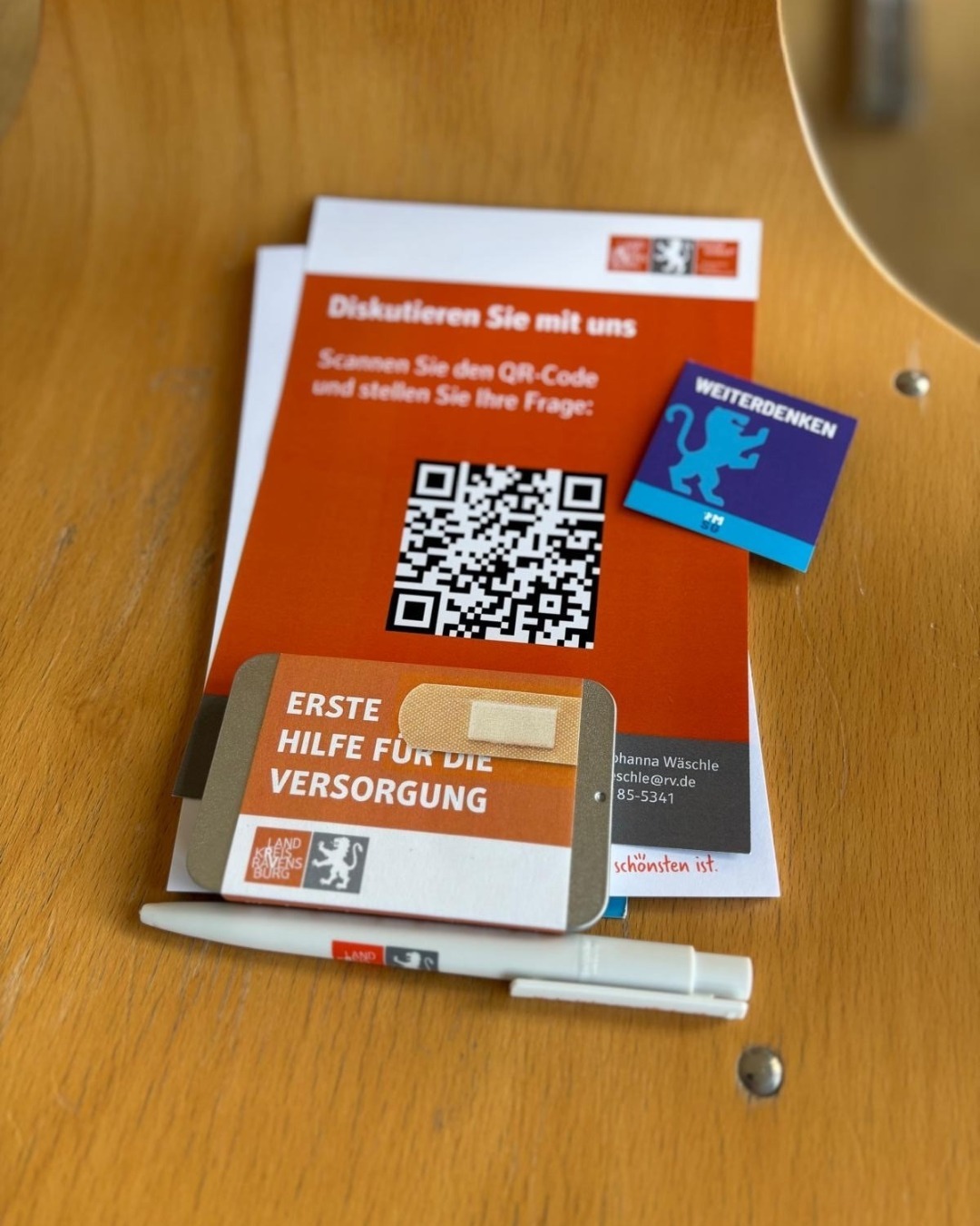 Geschenkartikel auf einem Stuhl, darunter eine Pflasterdose mit der Aufschrift: Erste Hilfe für die Versorgung.