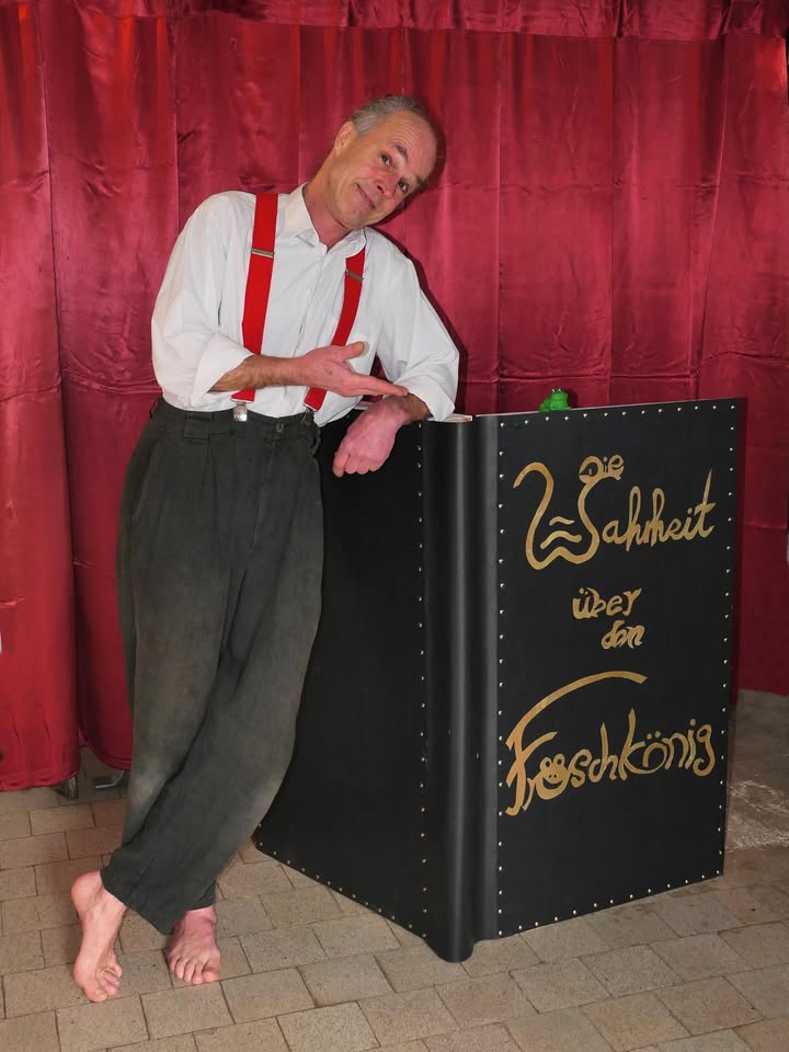 Dieser Alternativtext wurde KI-generiert: Ein Mensch in Hosenträgern lehnt an einem schwarzen, schmuck verzierten Podest. Im Hintergrund ist ein roter Vorhang zu sehen.