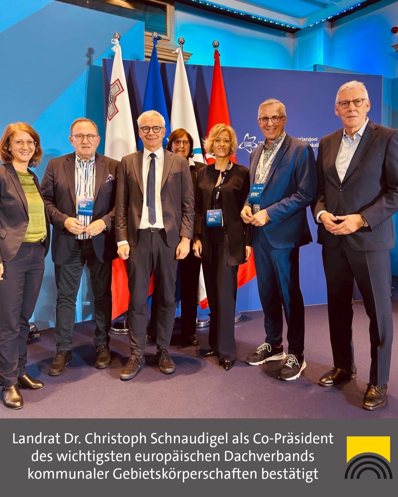 Dieser Alternativtext wurde KI-generiert: Eine farbenfrohe Aufnahme zeigt eine Gruppe von Personen vor mehreren Flaggen. Sie stehen auf einem Teppichboden und scheinen ein formelles Ereignis zu besuchen.