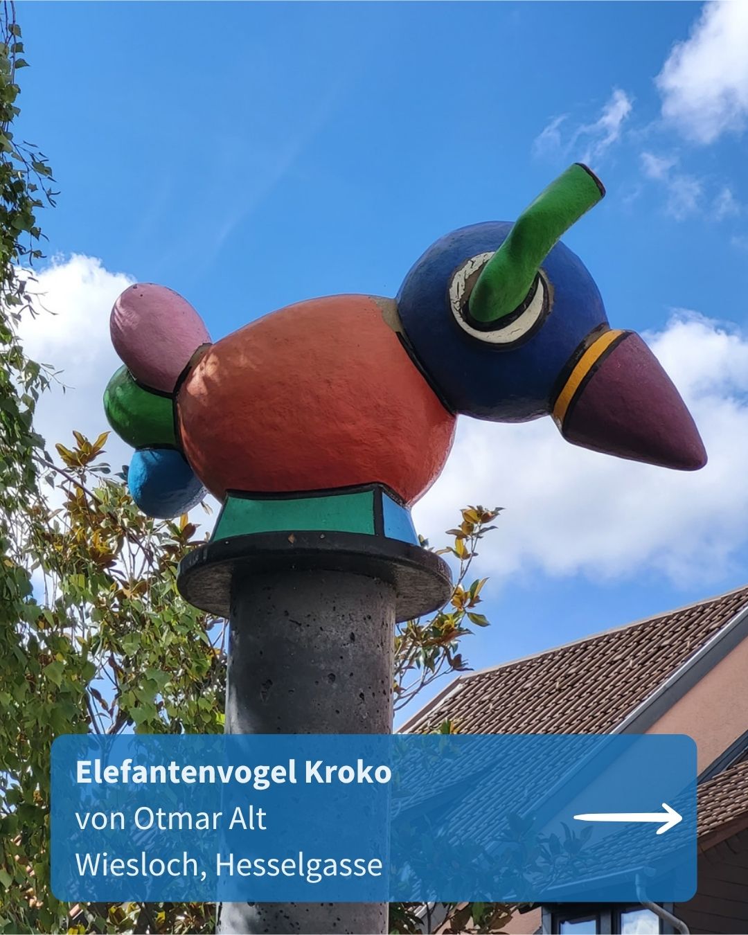 Farbenfrohe Skulptur eines Elefantenvogels mit blauem Kopf, grünem Horn und orangefarbenem Körper auf einem grauen Betonpfeiler vor blauem Himmel. Ein blauer Textkasten unten enthält die Aufschrift „Elefantenvogel Kroko von Otmar Alt Wiesloch, Hesselgasse“ und einen weißen Pfeil nach rechts. (KI-Alternativtext)