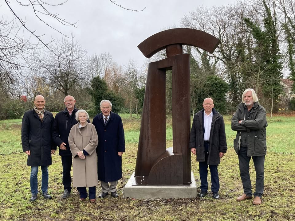 Dieser Alternativtext wurde KI-generiert: Eine Gruppe von Menschen steht vor einer modernen, braunen Skulptur im Freien. Im Hintergrund sind Bäume und Büsche zu sehen.