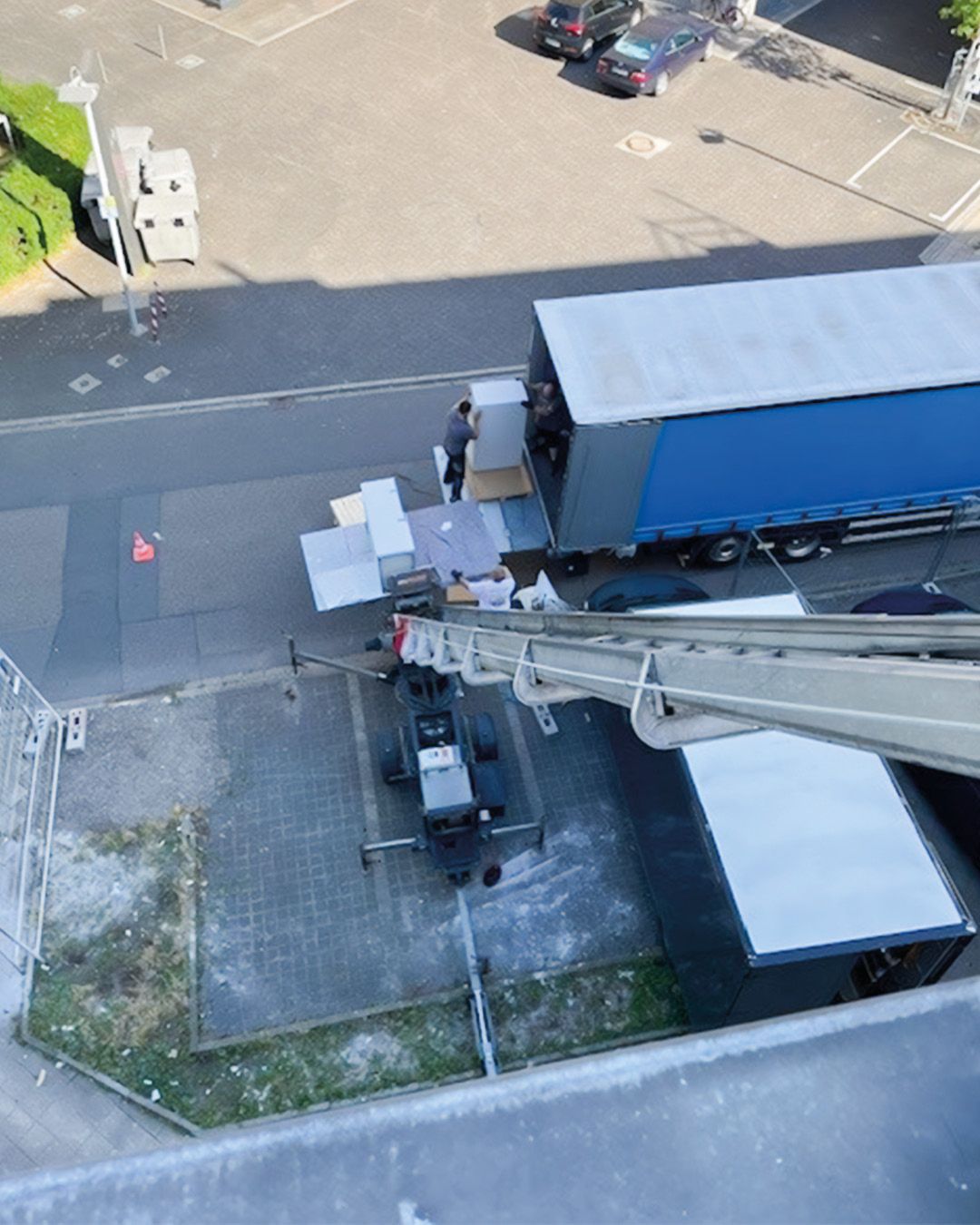 Dieser Alternativtext wurde KI-generiert: Eine Aufnahme von oben zeigt einen Lkw, der Gegenstände an ein Gebäude liefert. Ein Kran wird genutzt, um die Gegenstände hochzuheben.