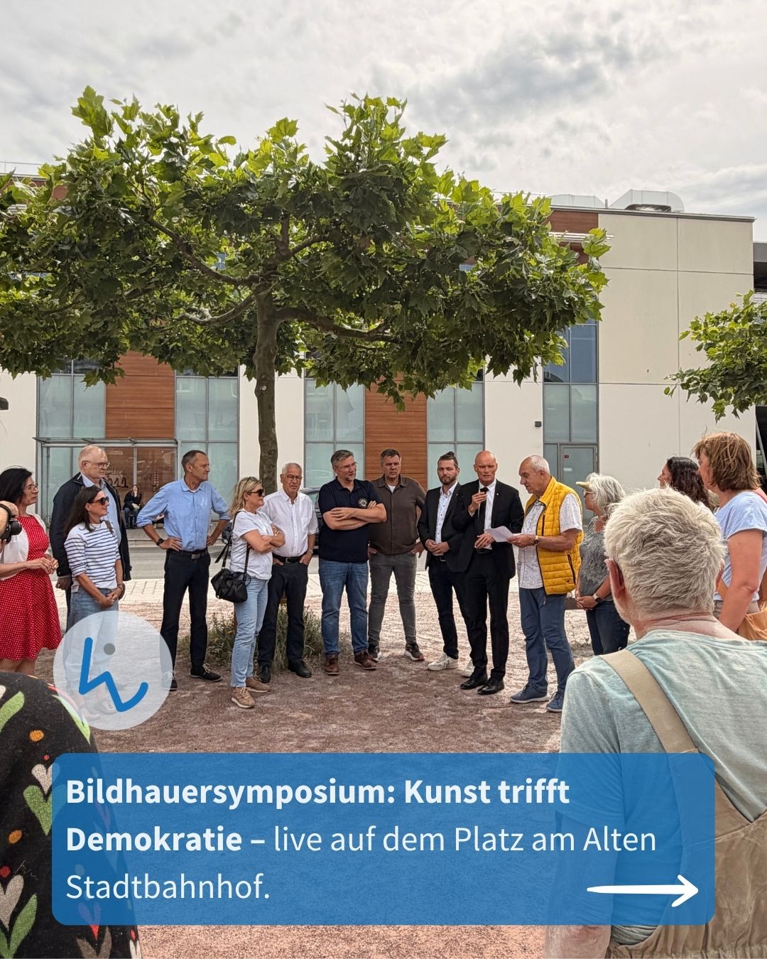 Gruppe von Menschen steht im Freien vor einem modernen Gebäude und hört einem Sprecher zu, der ein Blatt hält. Ein Baum steht im Vordergrund, ein blauer Textbalken unten mit der Aufschrift „Bildhauersymposium: Kunst trifft Demokratie – live auf dem Platz am Alten Stadtbahnhof“. (KI-Alternativtext)