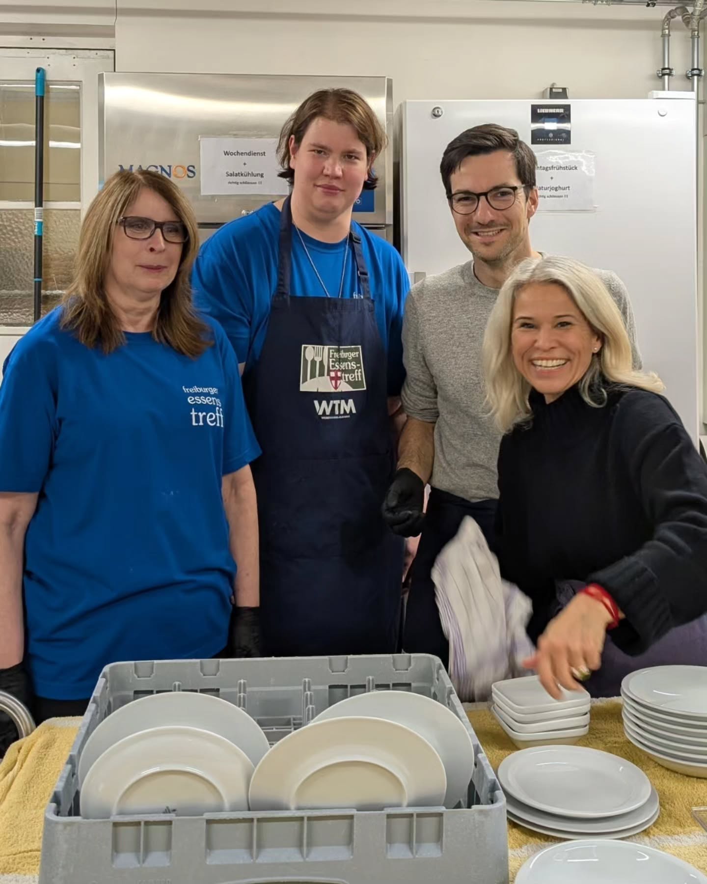 Dieser Alternativtext wurde KI-generiert: Eine Gruppe von Menschen steht in einem hellen Raum mit einer großen Metallausrüstung im Hintergrund. Sie halten Geschirr und scheinen gemeinsam zu arbeiten.