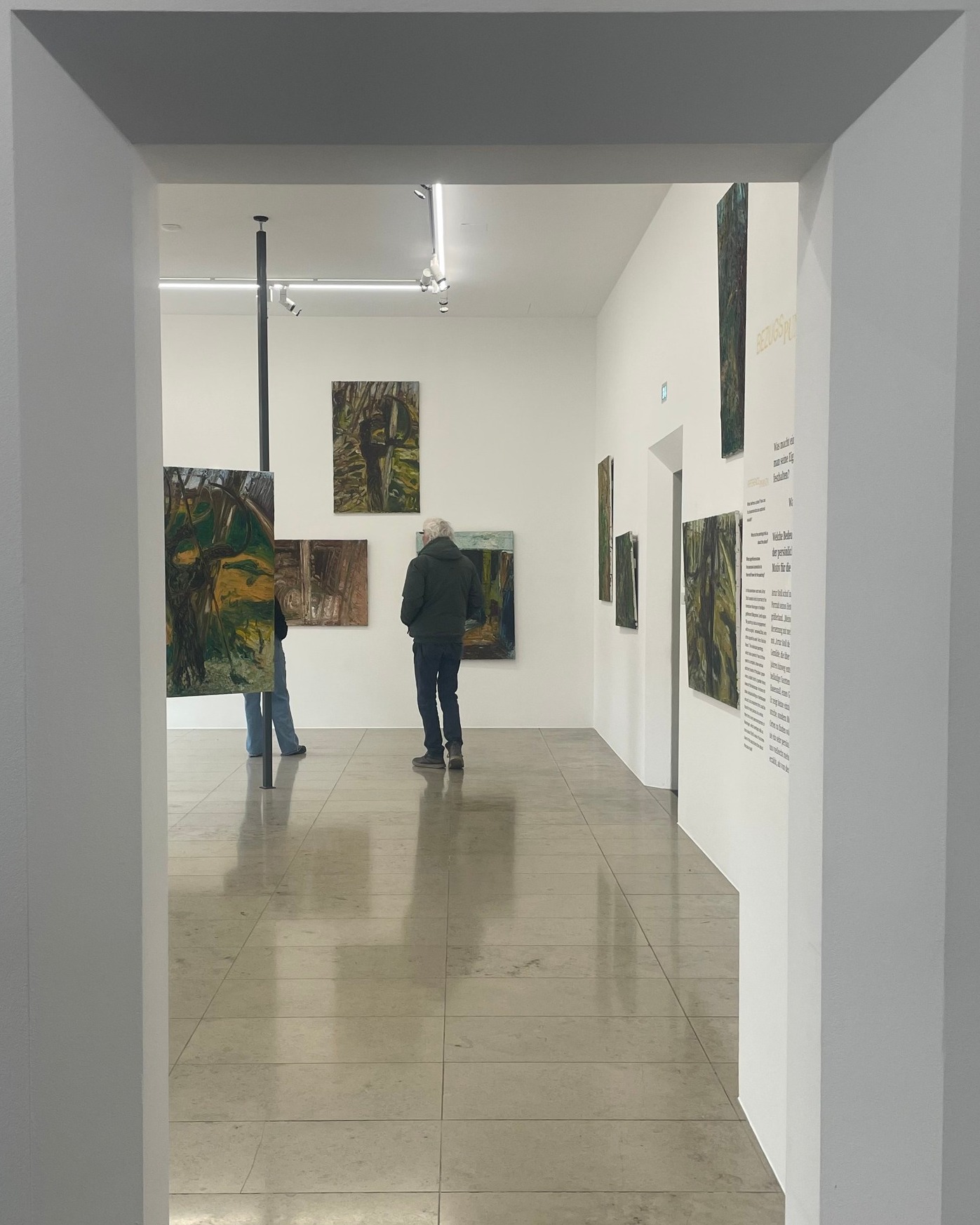 Dieser Alternativtext wurde KI-generiert: Ein Innenraum mit Kunstwerken an den Wänden. Eine Person steht mit dem Rücken zum Betrachter vor den Gemälden.