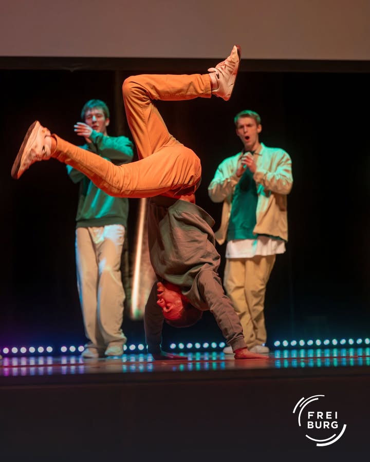 Ein Breakdancer zeigt eine Handstand-Bewegung auf einer Bühne, während zwei weitere Personen im Hintergrund stehen.  Die Szene ist beleuchtet.