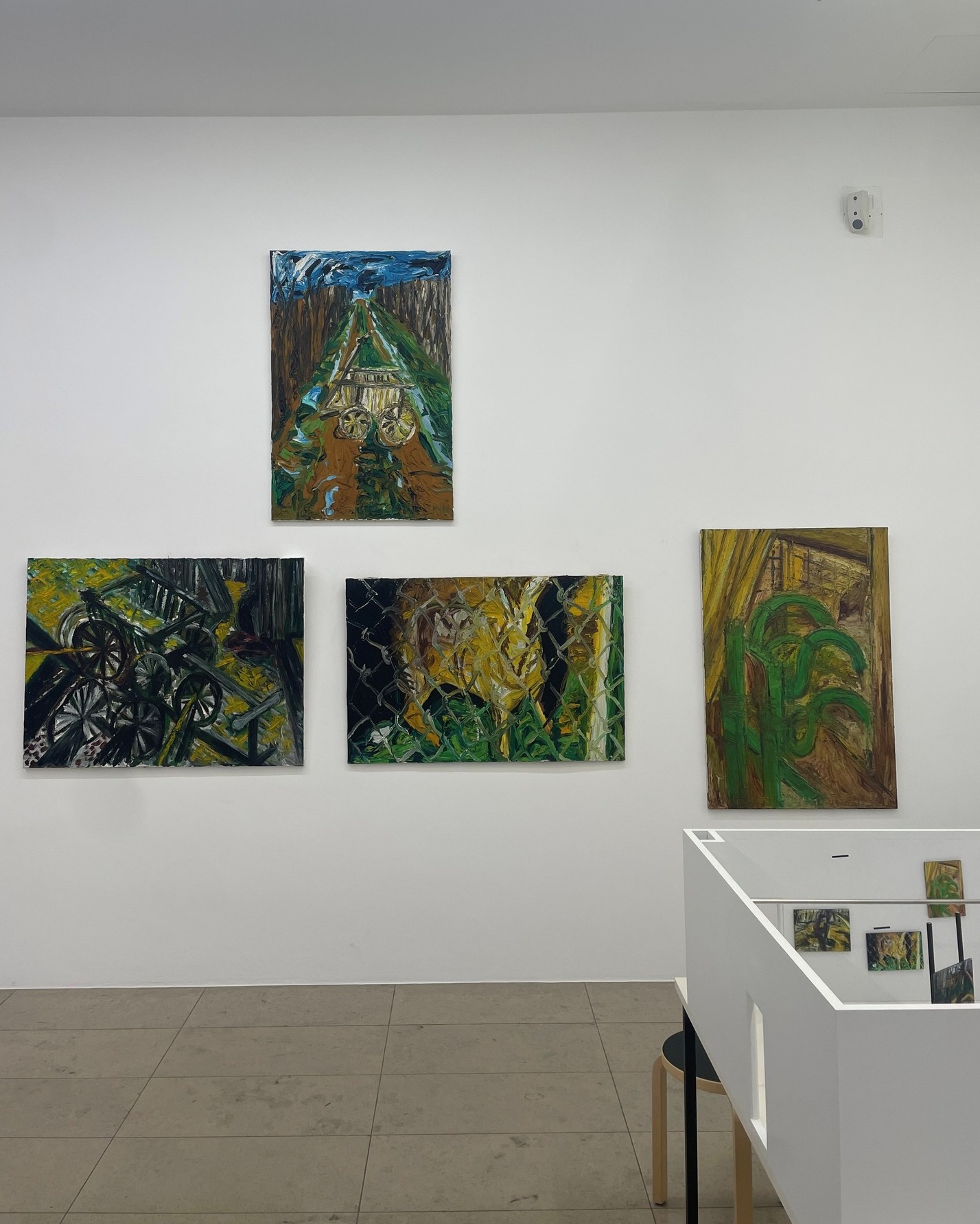 Dieser Alternativtext wurde KI-generiert: Mehrere farbenfrohe Gemälde hängen an einer hellen Wand in einem Raum. Im unteren Bildbereich ist ein Teil eines Tisches mit weiteren kleinen Bildern zu sehen.