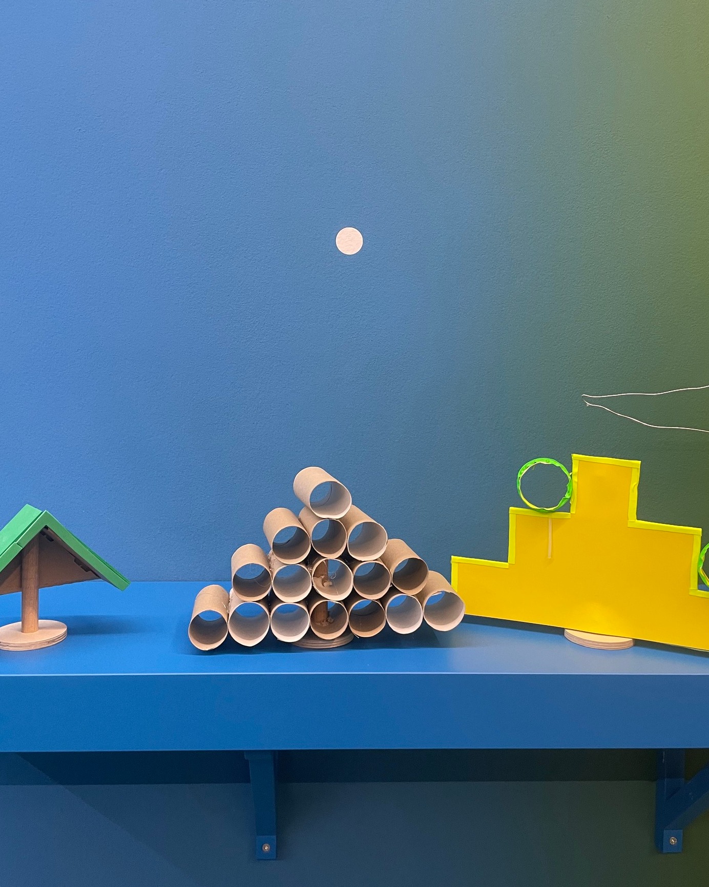 Auf einem blauen Regal vor einer blau-grün verlaufenden Wand stehen drei Objekte: links ein kleines Holzhaus mit grünem Dach, in der Mitte eine Pyramide aus Pappröhren und rechts eine gelbe Treppenstruktur mit einem grünen Ring. An der Wand darüber ist ein kleiner weißer Kreis angebracht. (KI-Alternativtext)