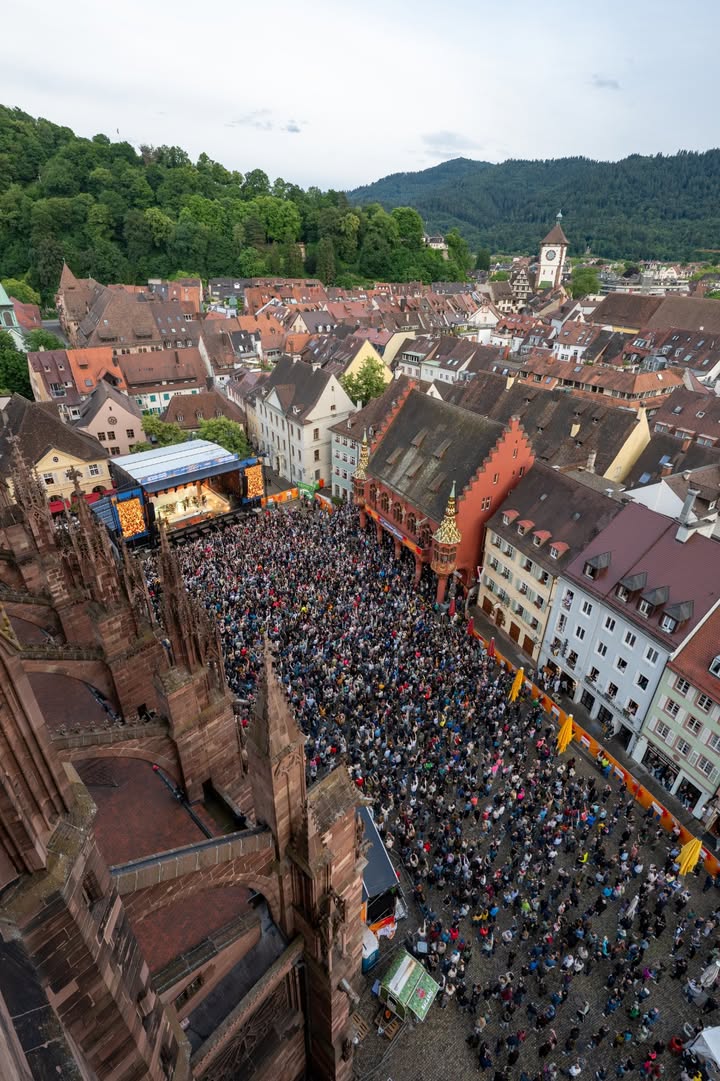 Große Menschenmenge auf einem Marktplatz vor historischen Gebäuden und einer gotischen Kirche, im Hintergrund bewaldete Hügel. Ein Bühnenpodest mit Beleuchtung ist aufgebaut, gelbe Markisen säumen den Platz. (KI-Alternativtext)