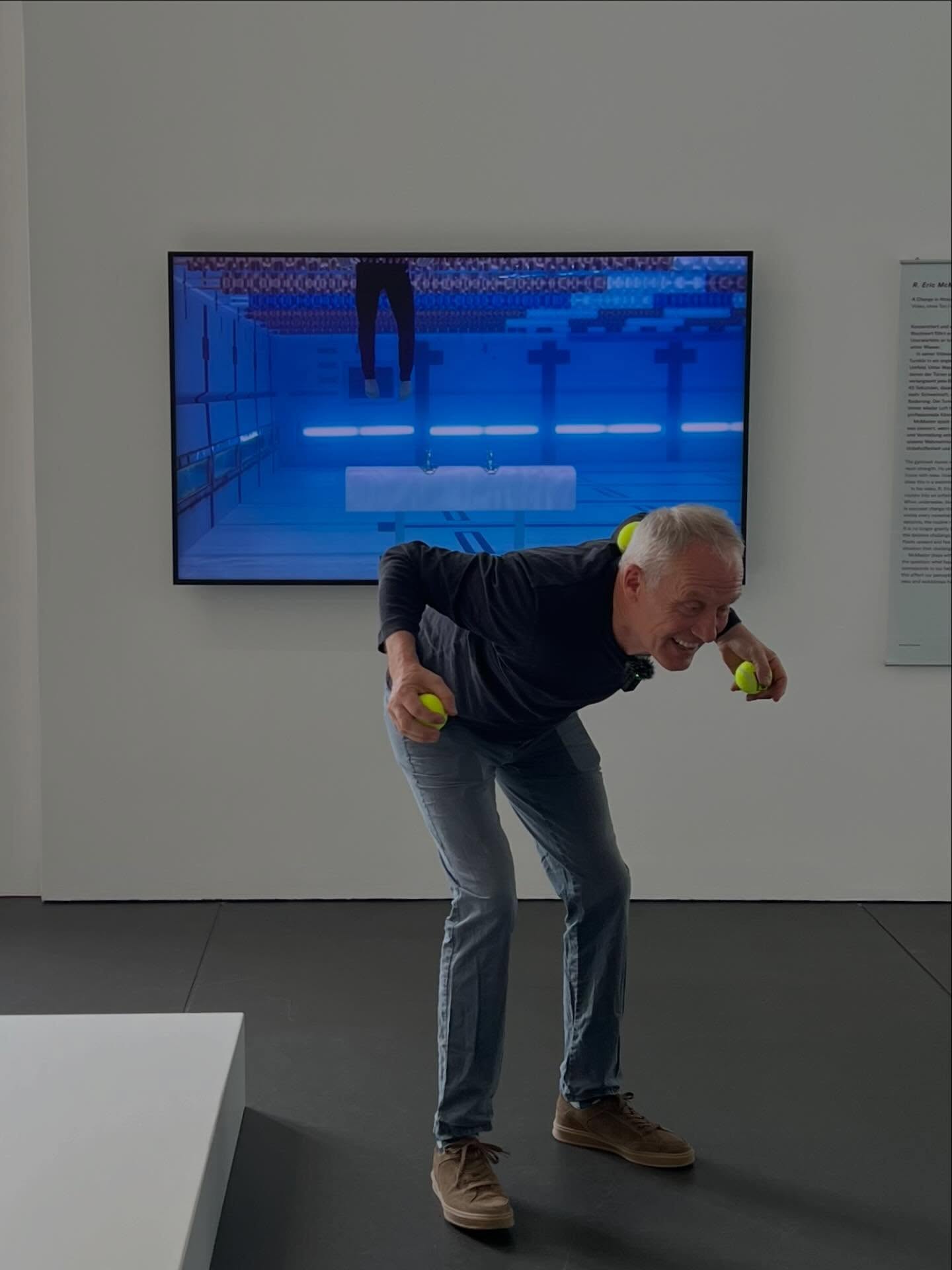 Dieser Alternativtext wurde KI-generiert: Eine Person steht vor einem Bildschirm, der ein Tennisfeld zeigt. Die Person hält zwei gelbe Bälle hoch und scheint sich zu bewegen.