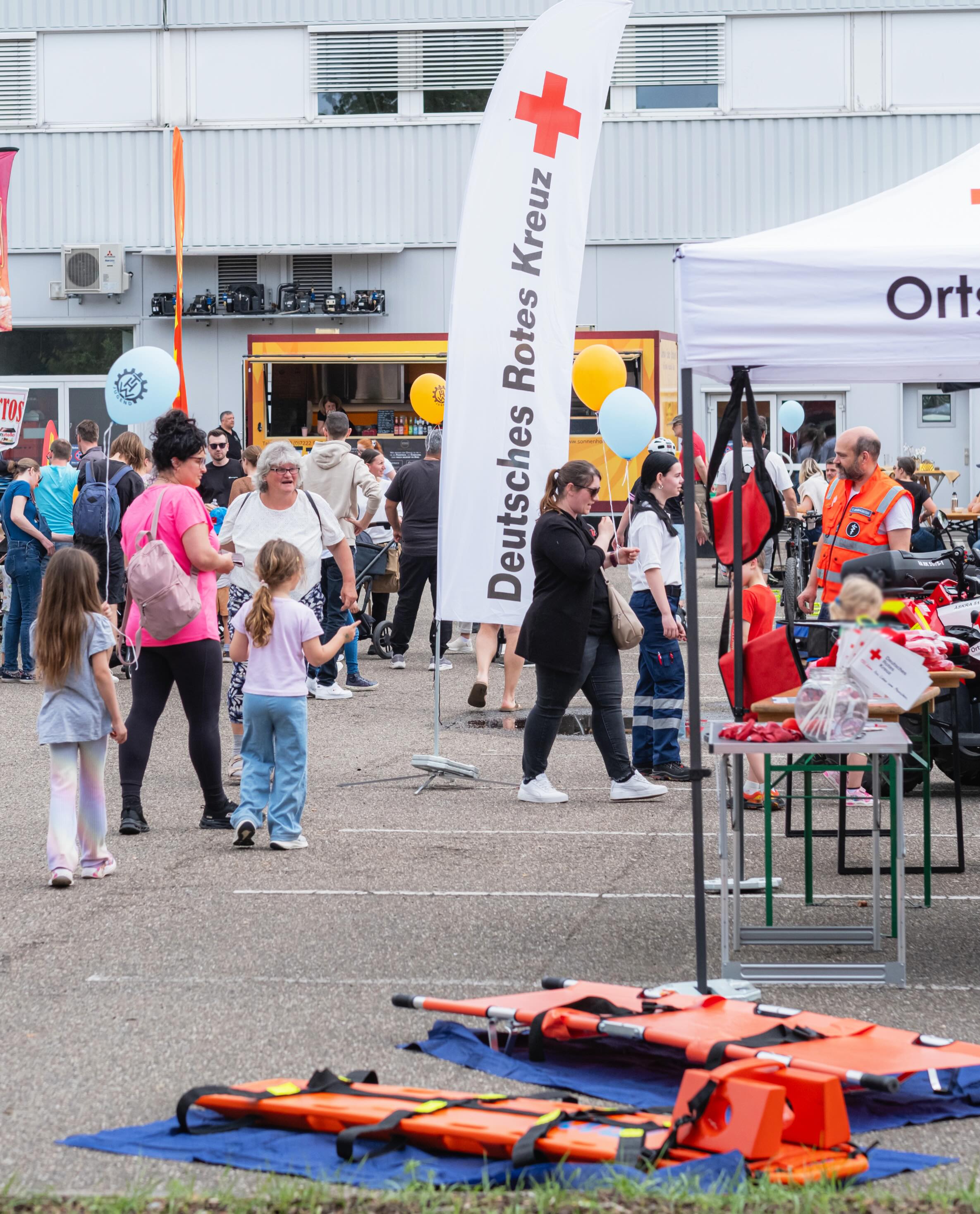 Dieser Alternativtext wurde KI-generiert: Eine Menschengruppe steht vor einem Gebäude und einem Zelt mit dem Zeichen eines Rettungsdienstes. Im Vordergrund liegen auf einer blauen Decke orangefarbene Tragen.